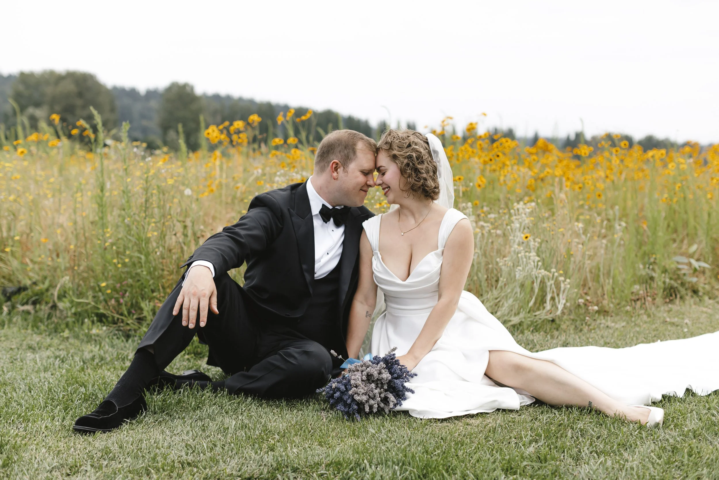 A newlywed couple sits on grass in a field of yellow flowers, touching foreheads and smiling, with the bride holding a bouquet of lavender.