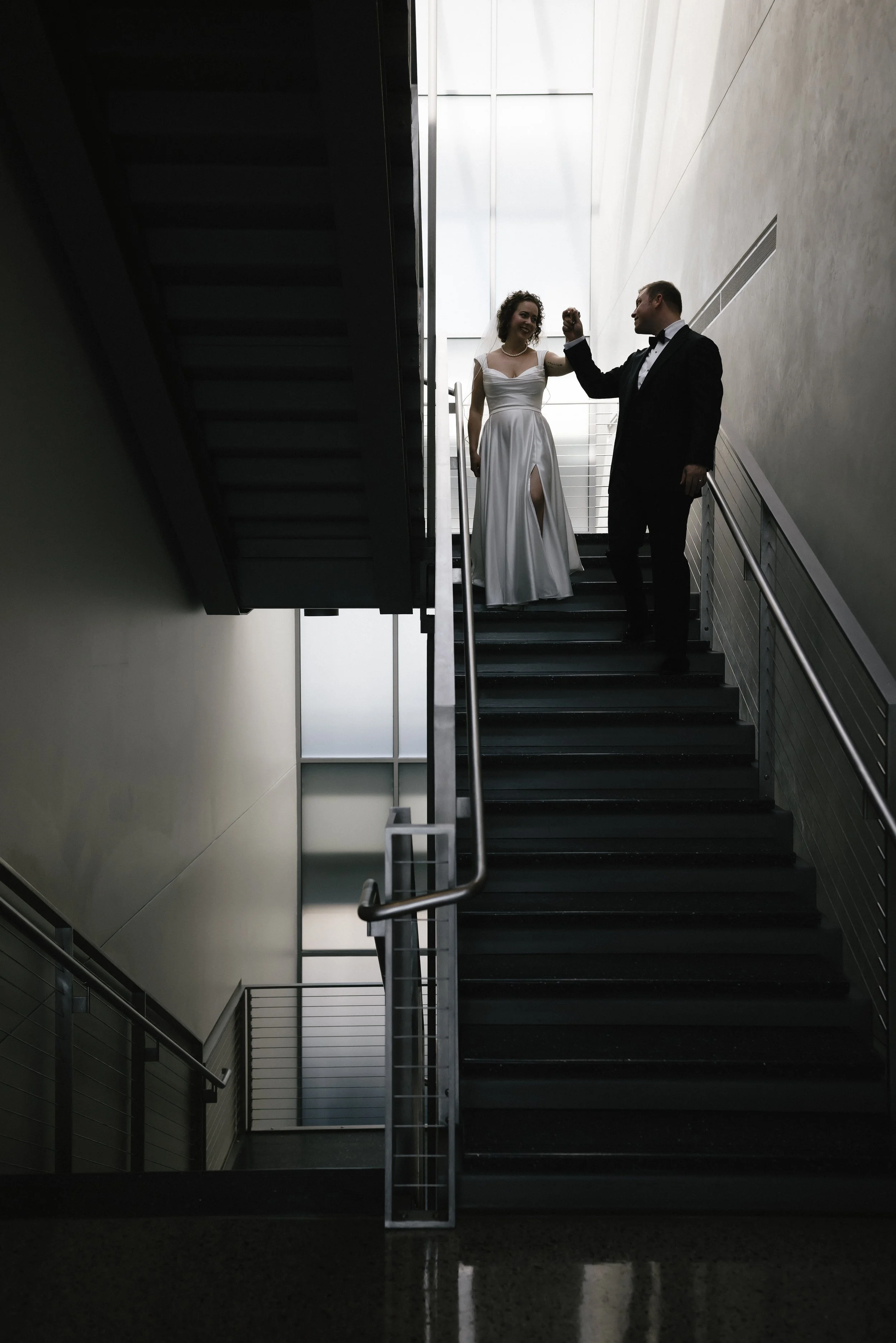 A bride and groom standing on a staircase, smiling and holding hands.