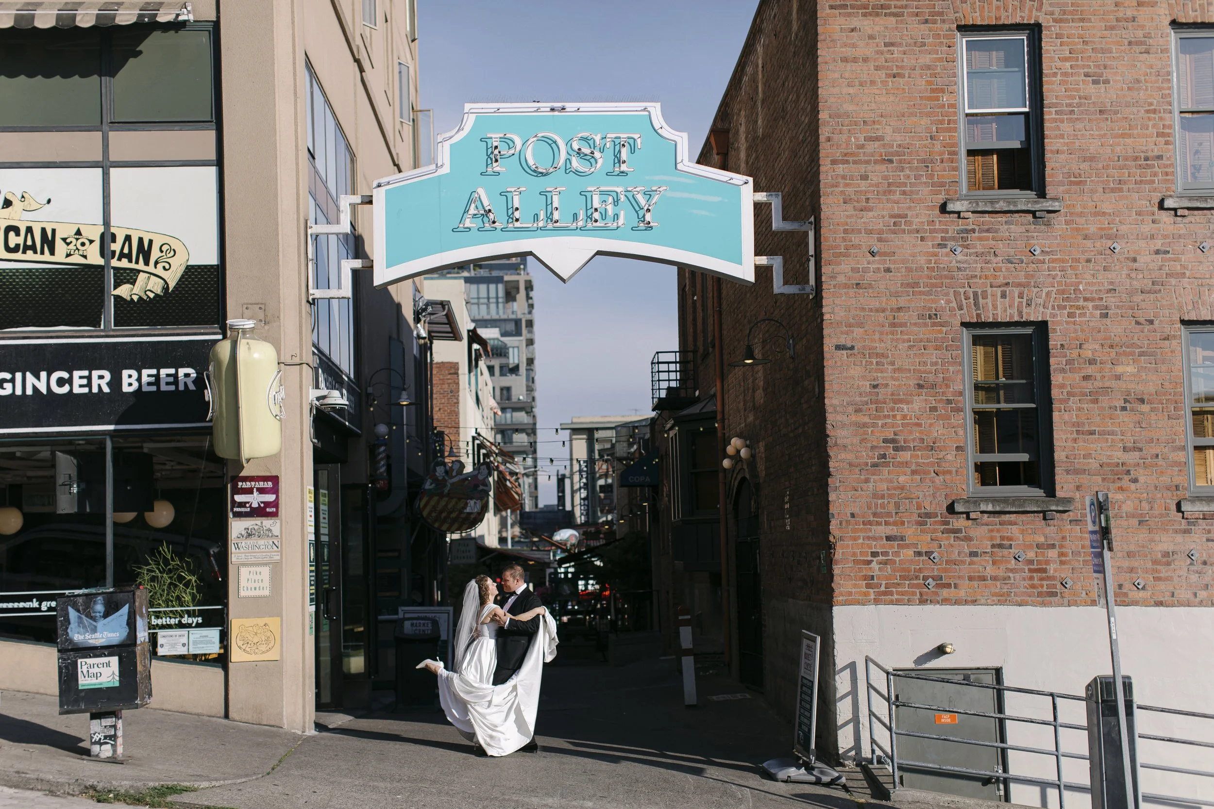 A bride and groom dancing in front of Post Alley, with the bride lifting her gown slightly, in an urban setting with brick and modern buildings around.