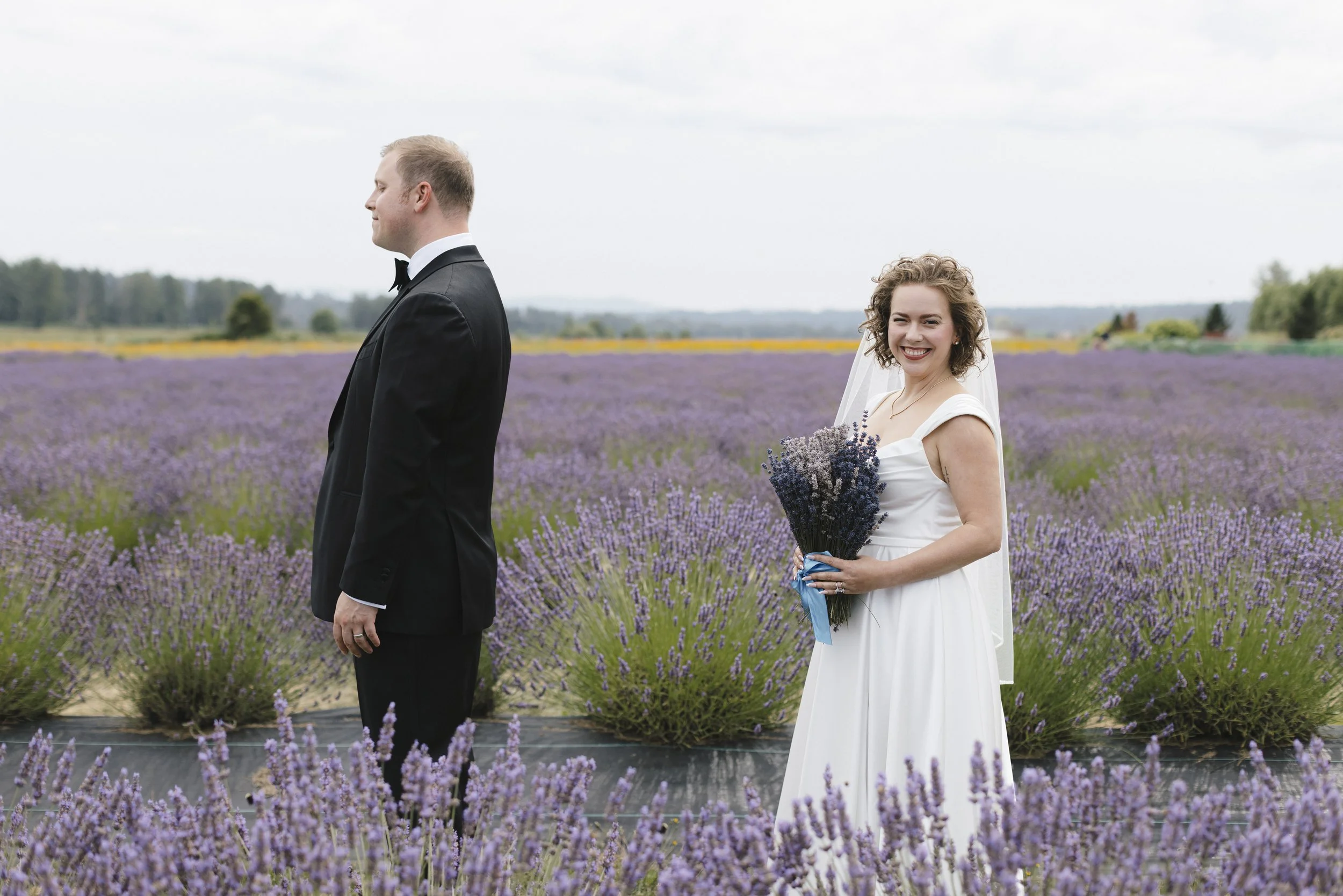 A bride in a white wedding dress holding a bouquet of lavender, smiling at the camera, standing in a lavender field, with a groom in a black suit and bow tie, facing away.