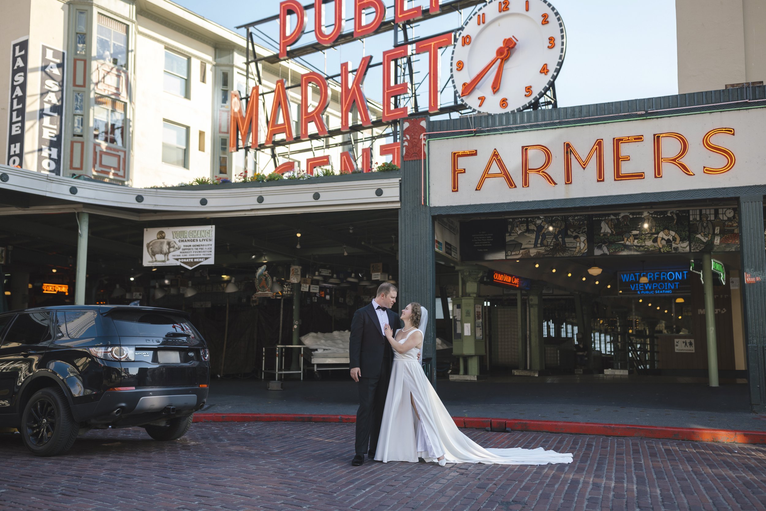 A newlywed couple in wedding attire standing on a brick street in front of Pike Place Market in Seattle, Washington, with neon signs and a large clock overhead.