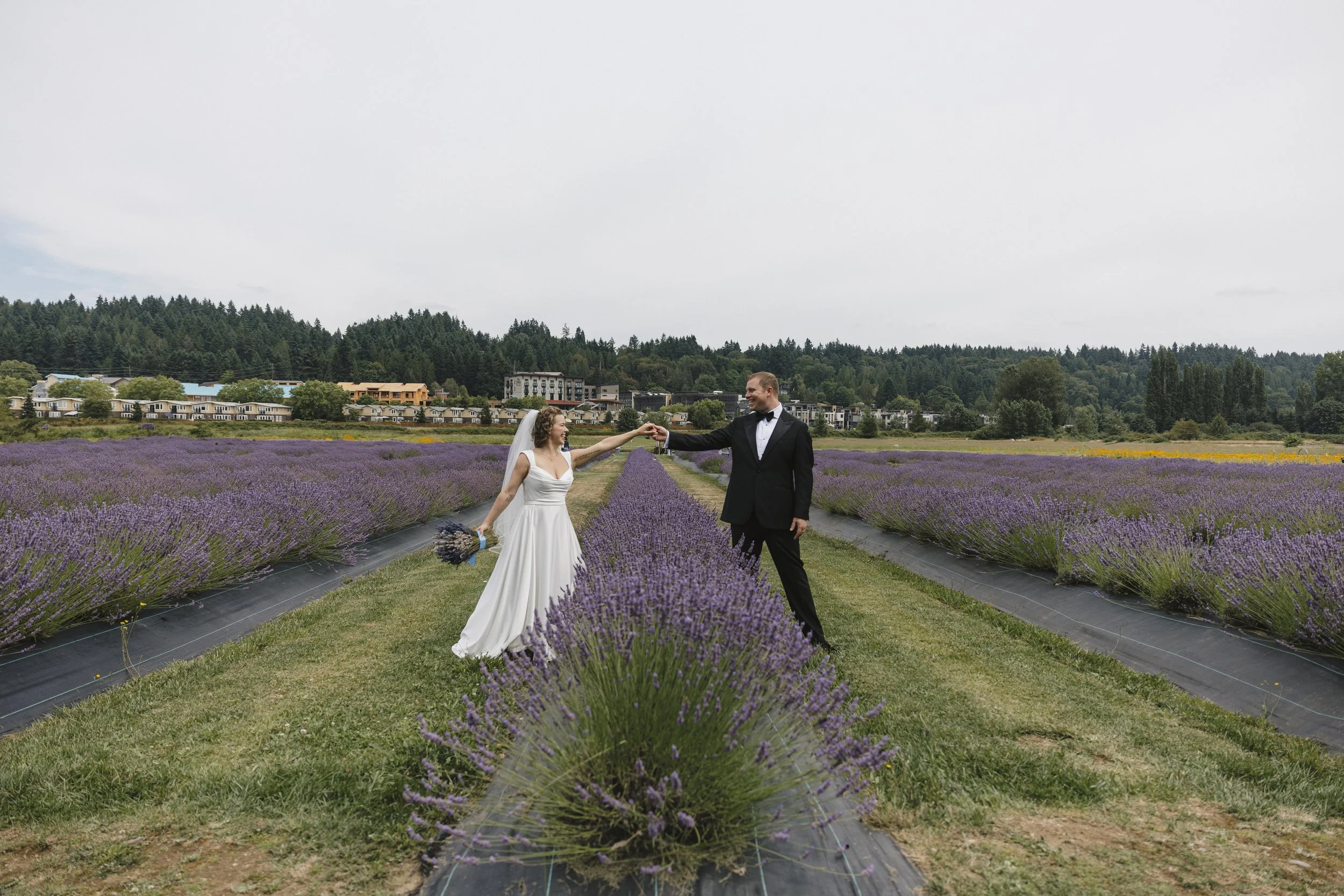 Bride and groom in wedding attire holding hands in lavender field.
