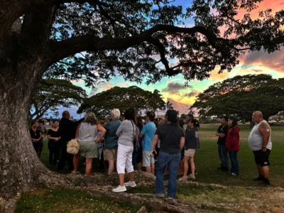 A group of people stand beneath a tree during a colorful sunset on the Waikiki Night Marchers ghost tour