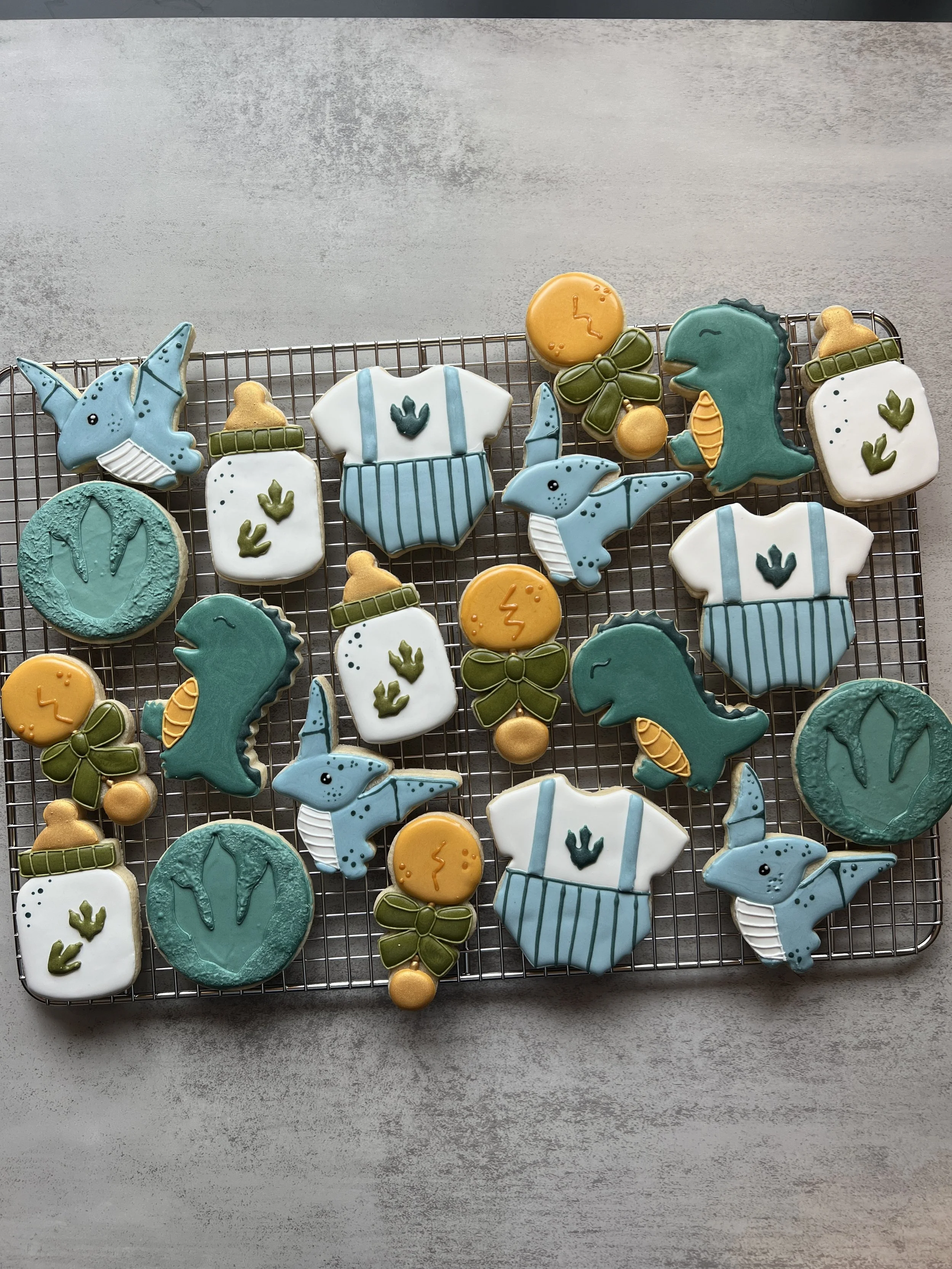 Assorted dinosaur-themed sugar cookies on a wire rack. The cookies include t-rex, pterodactyls, fossil footprint, baby clothes,  baby bottles, and rattles, decorated in colors of green, blue, while and dark yellow.