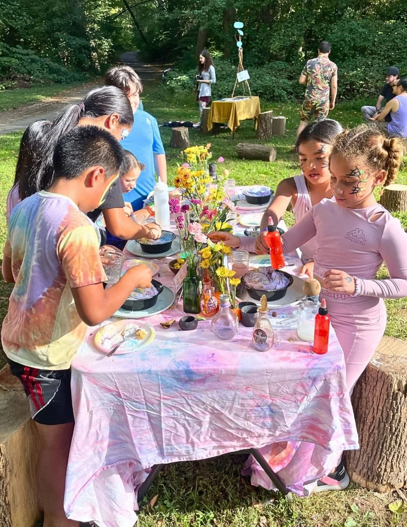 Children participating in a colorful outdoor craft activity at a forest birthday party, with a decorated table, flowers, and face paint, in a wooded setting.
