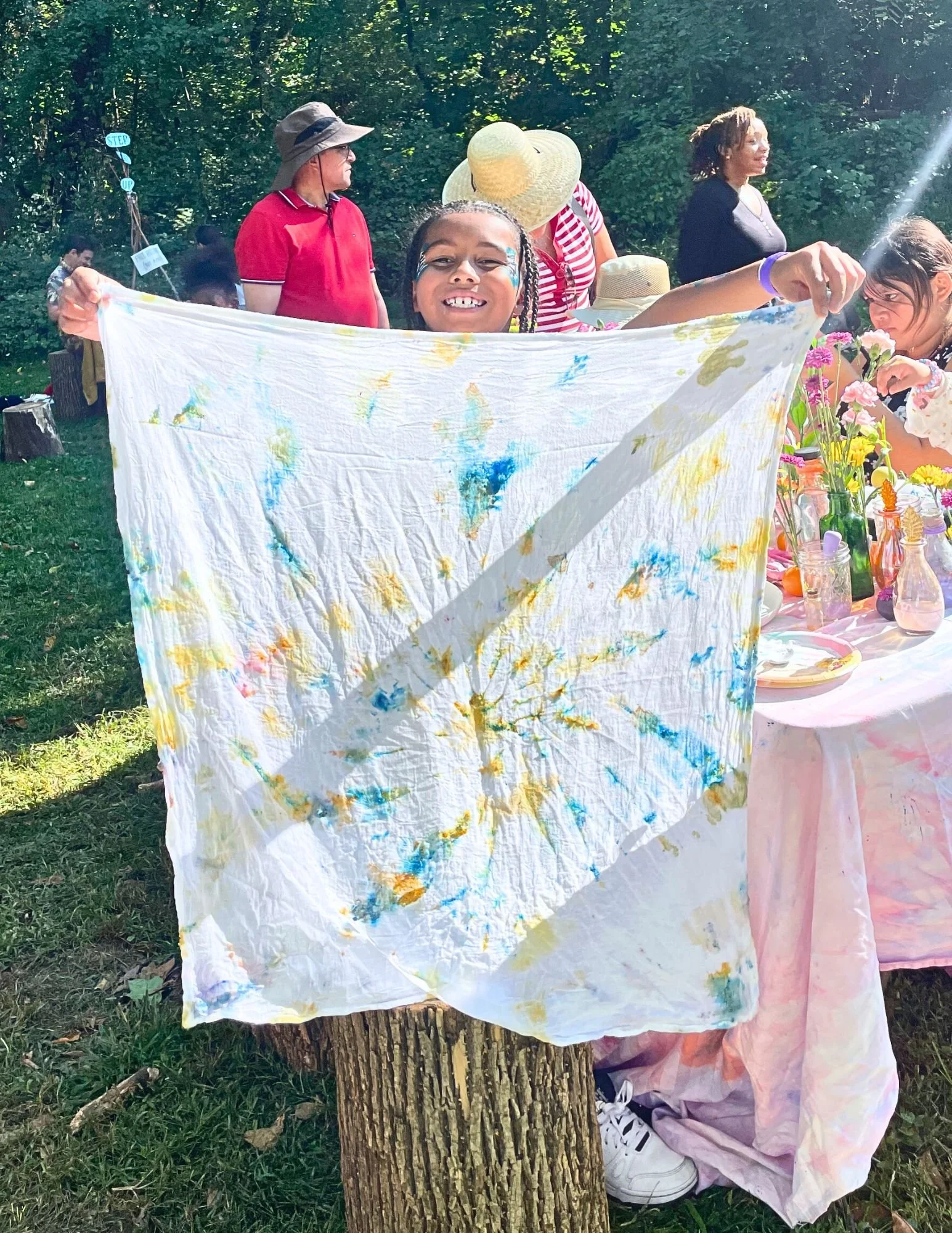 A young girl smiling and holding up a tie-dye fabric outdoors at a gathering. Several people, including women in hats, are mingling in the background, near a decorated table with flowers and bottles.