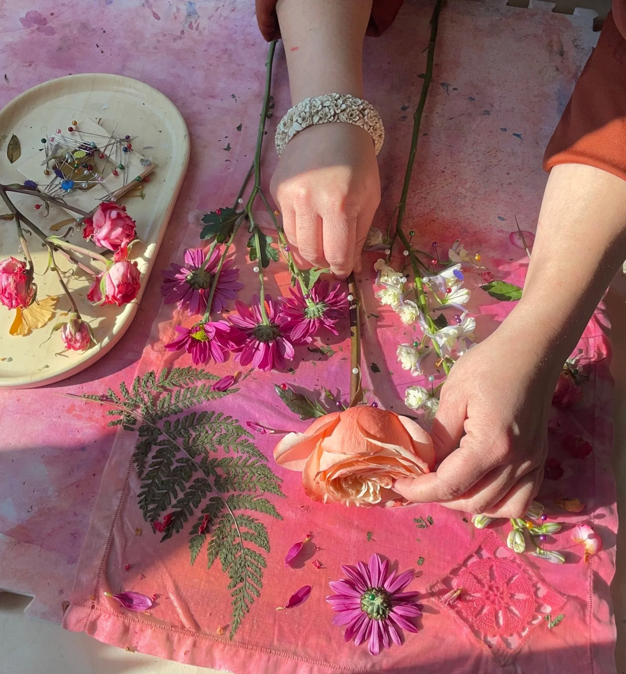 Person arranging pink, purple, white flowers and green fern on pink fabric, with accessories like pins on a tray nearby.