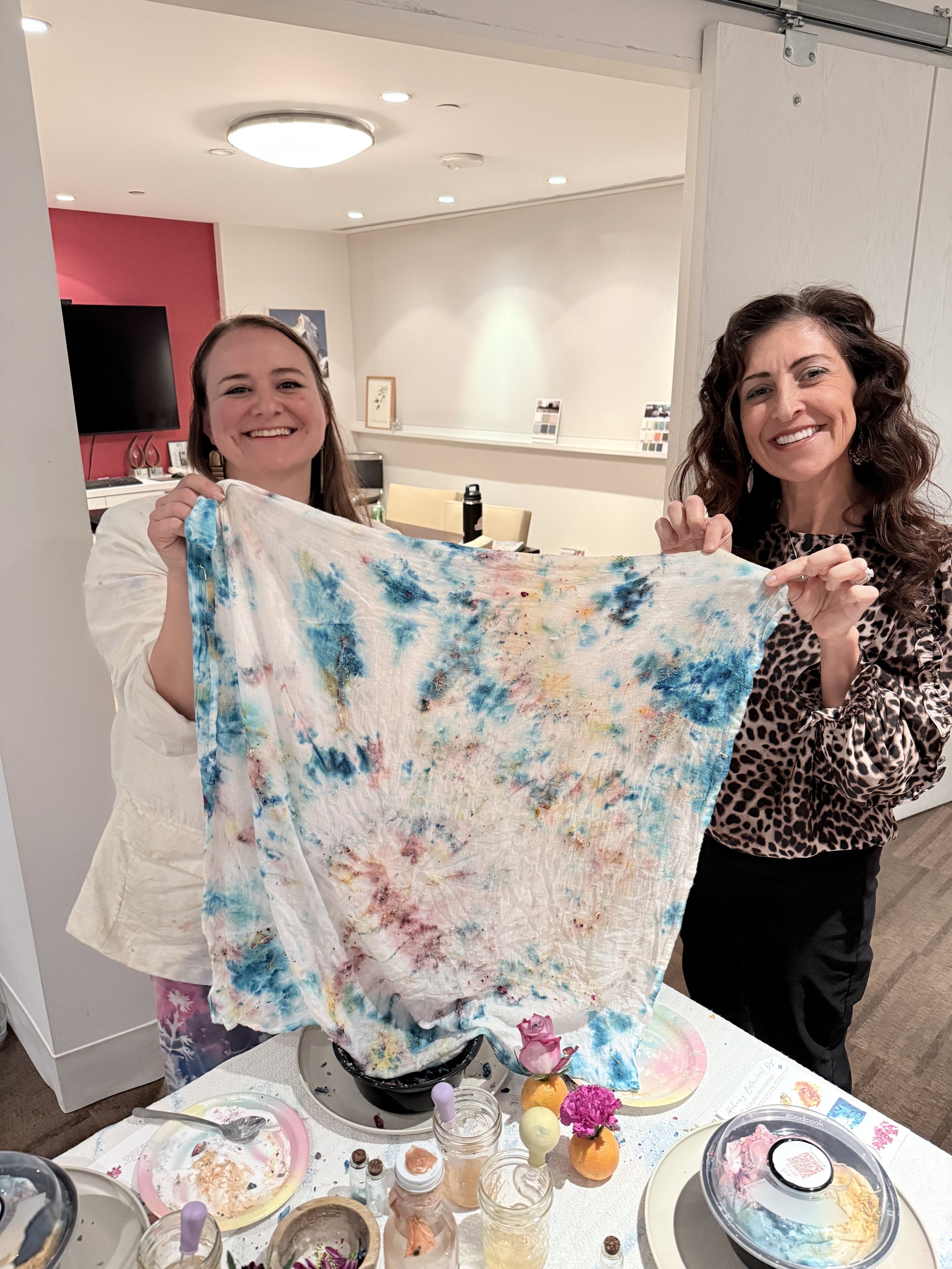 Two women smiling and holding a colorful tie-dye cloth in a home or art studio setting with art supplies on the table in front of them.