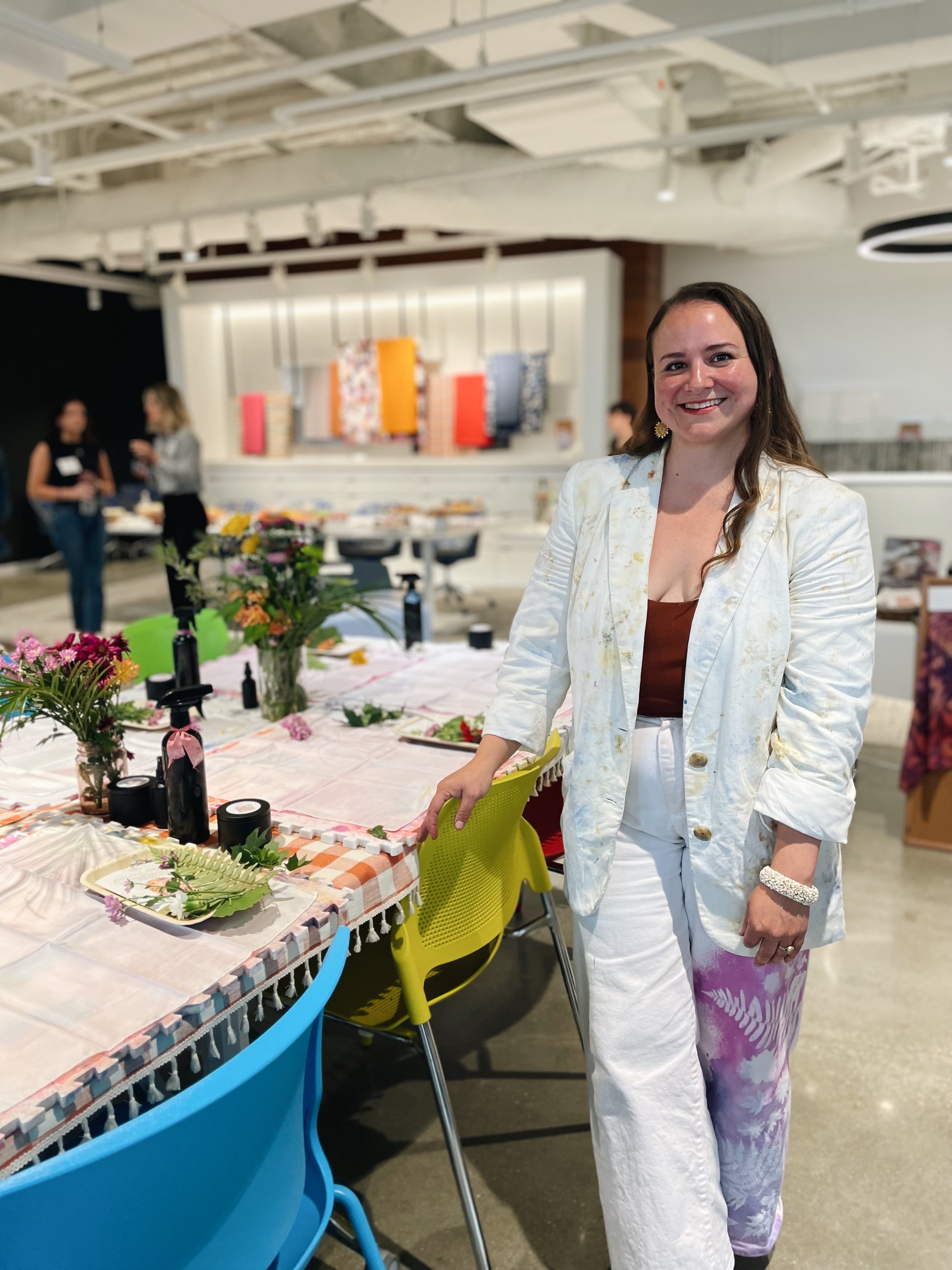 A woman standing beside a decorated table with floral arrangements and craft supplies in an art or event space.
