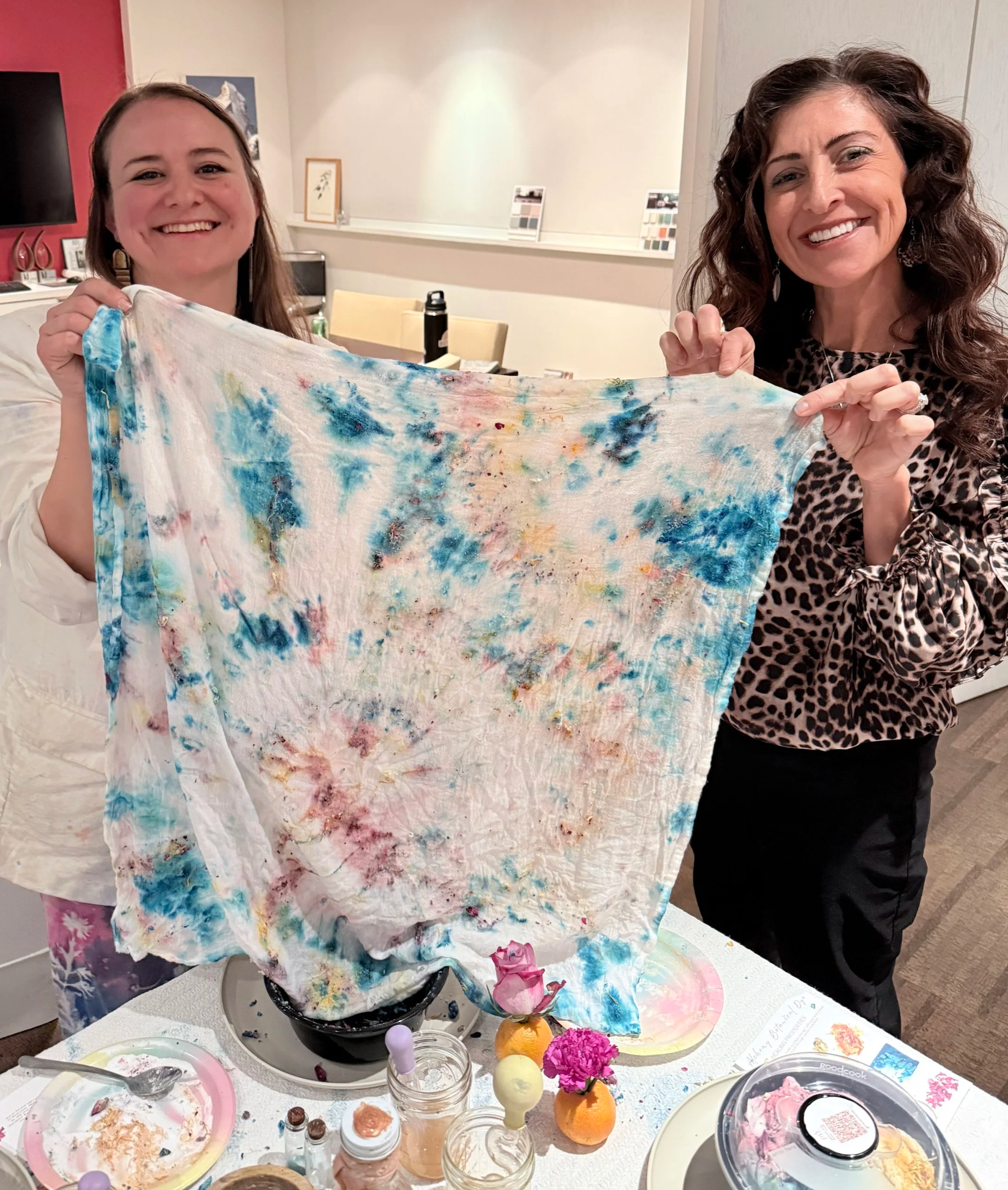 Two women holding a colorful tie-dye cloth in a room with art supplies and decorated table in front.