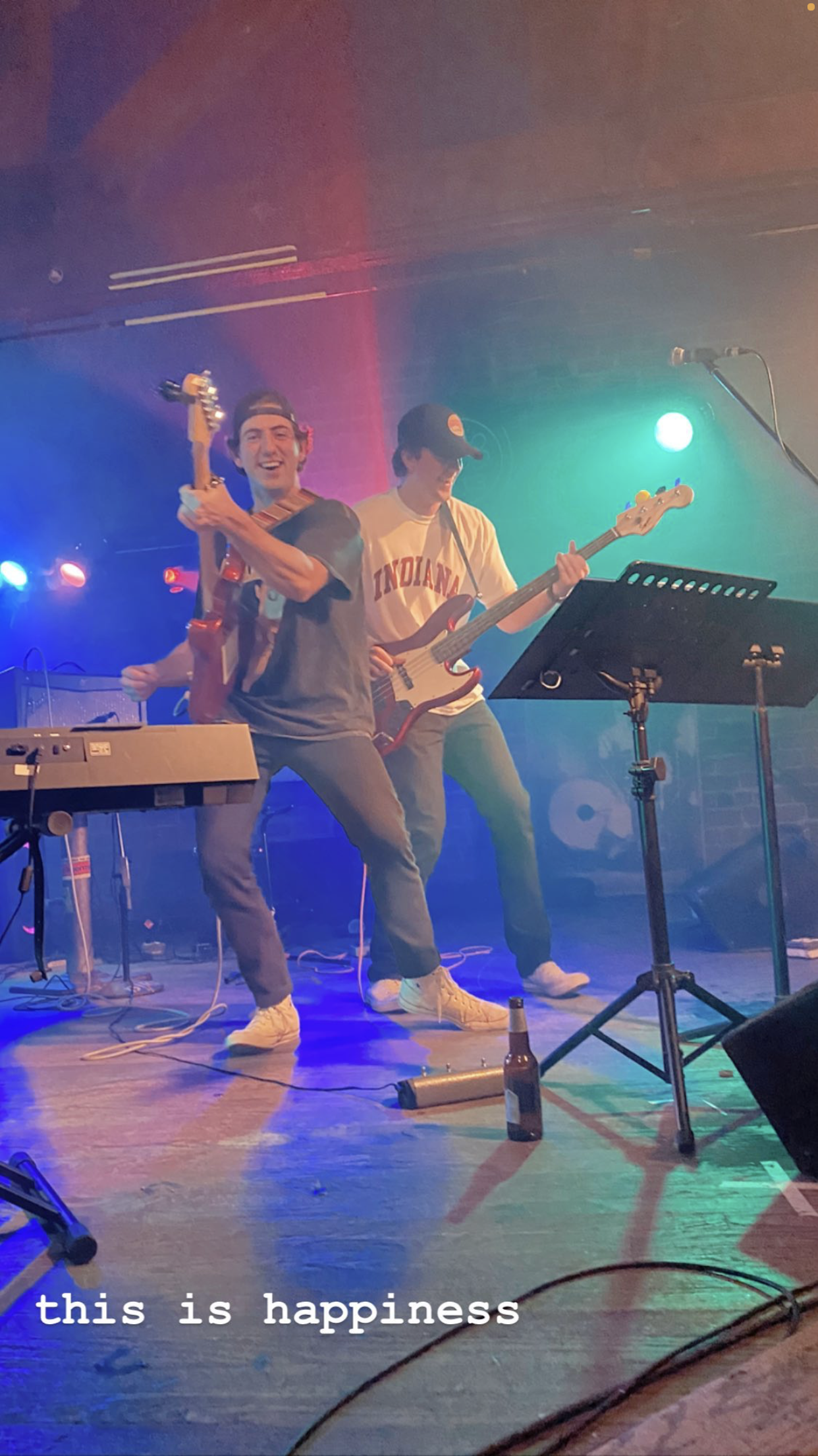 Two young men playing musical instruments on stage with colorful lights, one with a guitar and the other with a bass guitar, smiling and enjoying a live performance, with a microphone stand and a beer bottle on the floor.