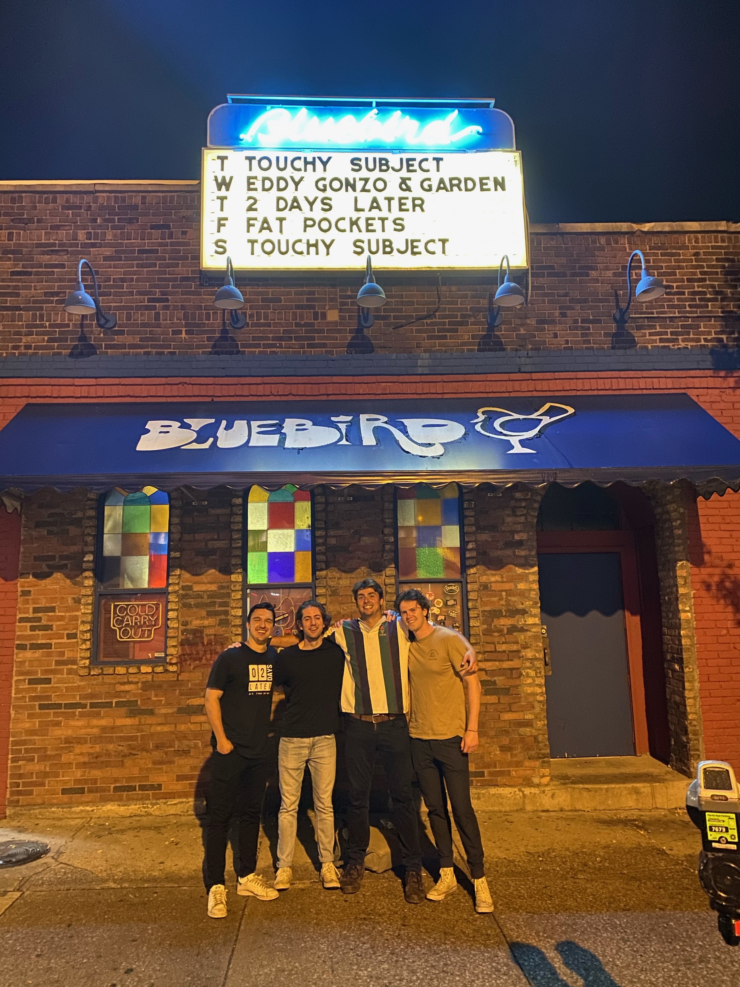 Four young men standing outside a brick building with stained glass windows and a blue awning, smiling at night.