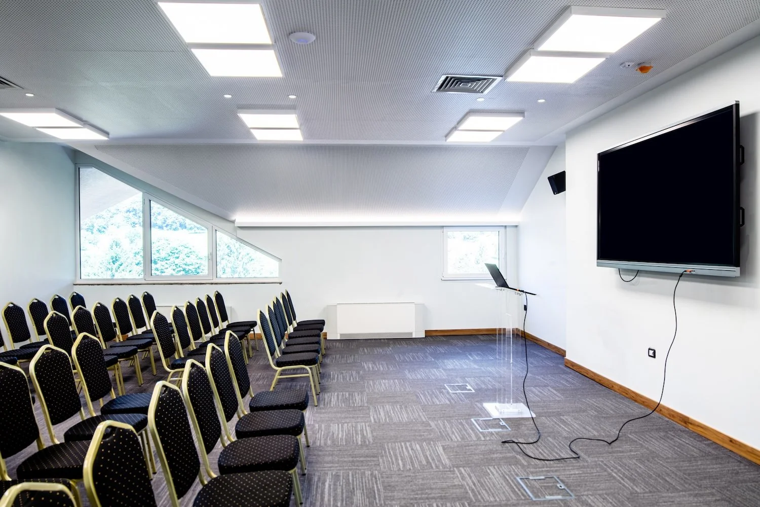 Empty conference room with rows of black chairs with gold trim, large window with a view of trees, a wall-mounted TV screen, and a laptop on a transparent stand  | Stay Cool ACR