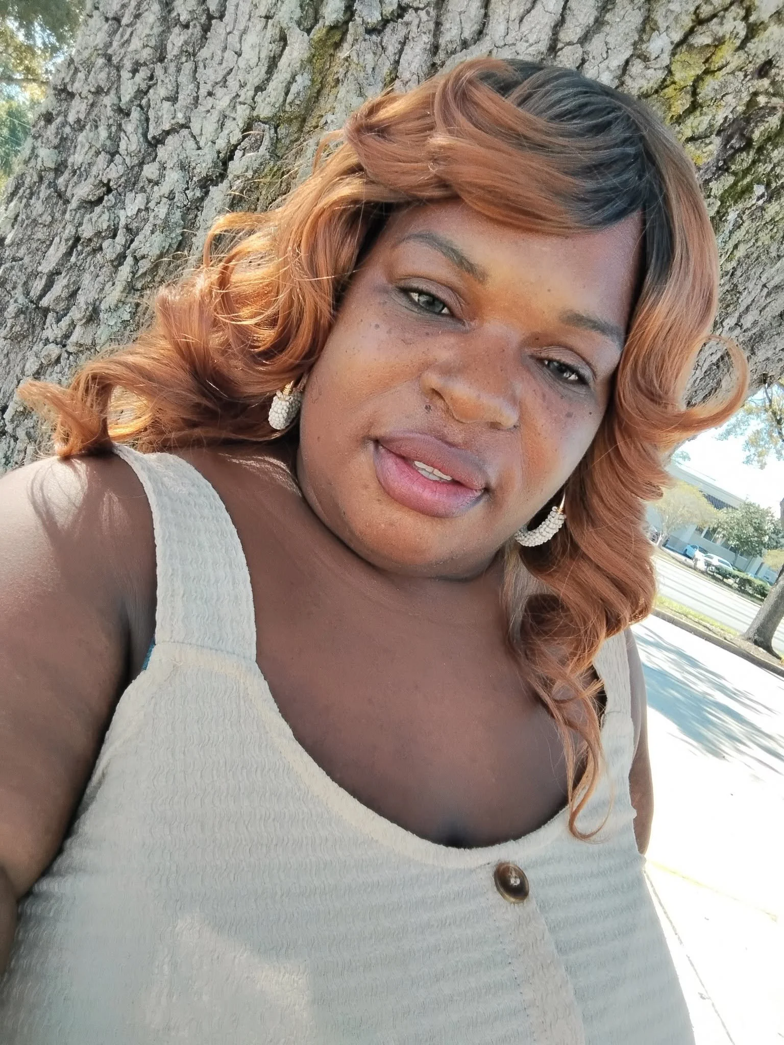 Close-up of a woman with brown hair styled in loose curls, wearing earrings and a light-colored top, standing outdoors next to a tree with a textured bark.