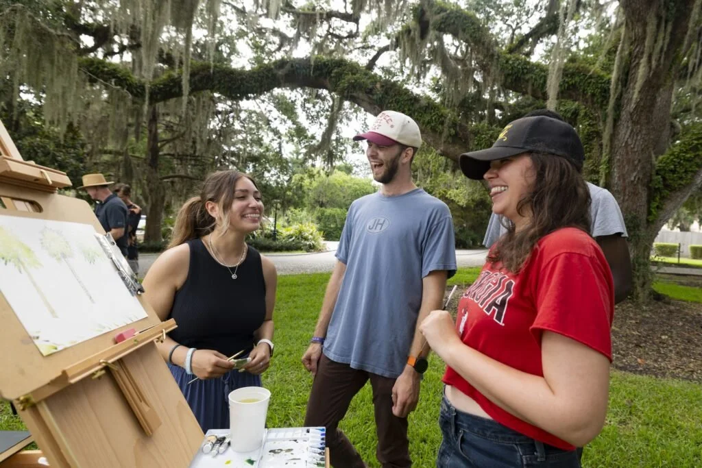 A group of college students talk and laugh as they paint outside on easels.