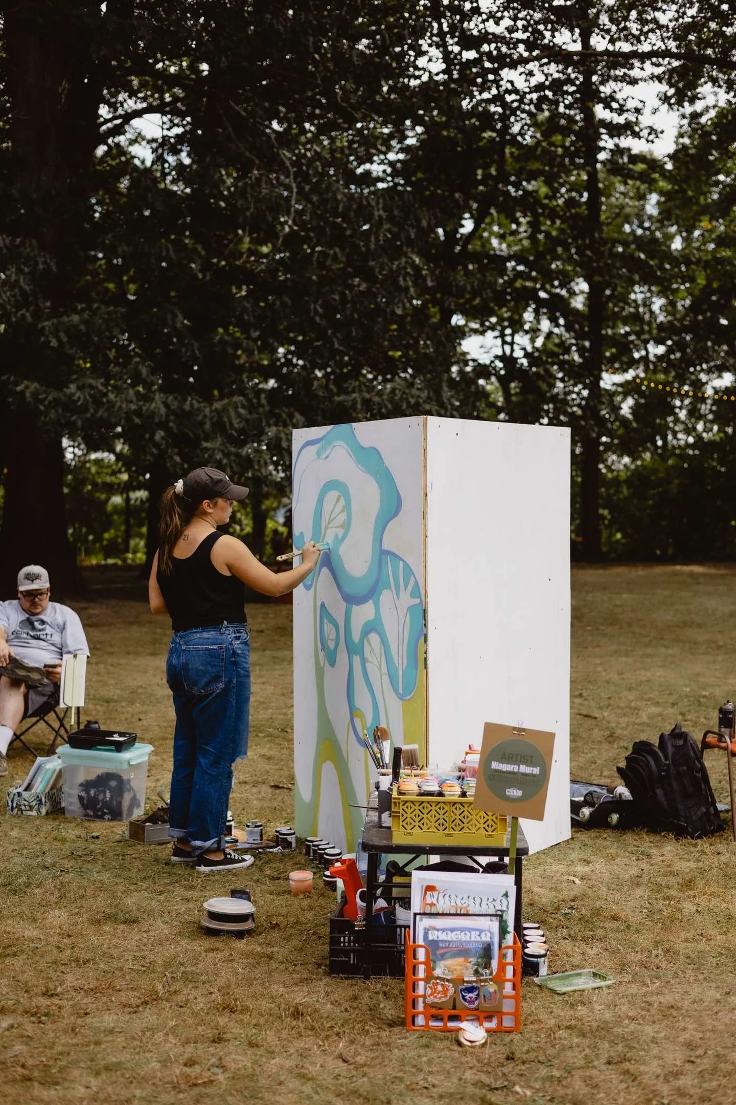 Artist painting abstract mural outdoors surrounded by art supplies, with a person sitting nearby in a park setting.