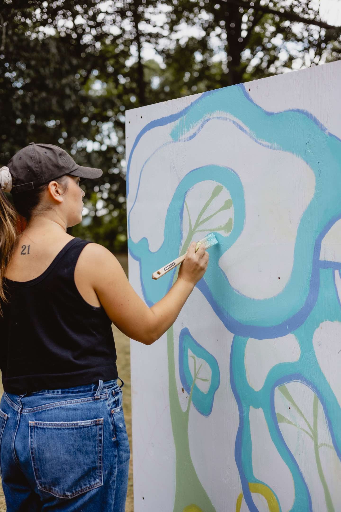 A person with a tattooed number 21 is painting a large abstract design with blue and green shapes on a board. They are outdoors, wearing a black tank top, blue jeans, and a black cap.