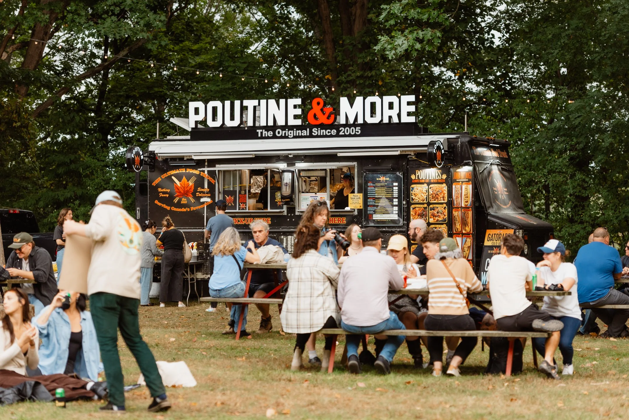 People sitting at picnic tables near a parked food truck labeled 'Poutine & More' in an outdoor setting with trees.