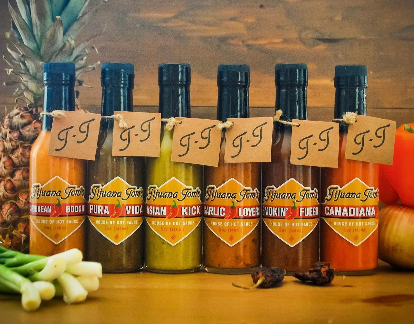 Assorted Tijuana Tom's hot sauce bottles with various flavors, surrounded by fresh and dried peppers and a pineapple.