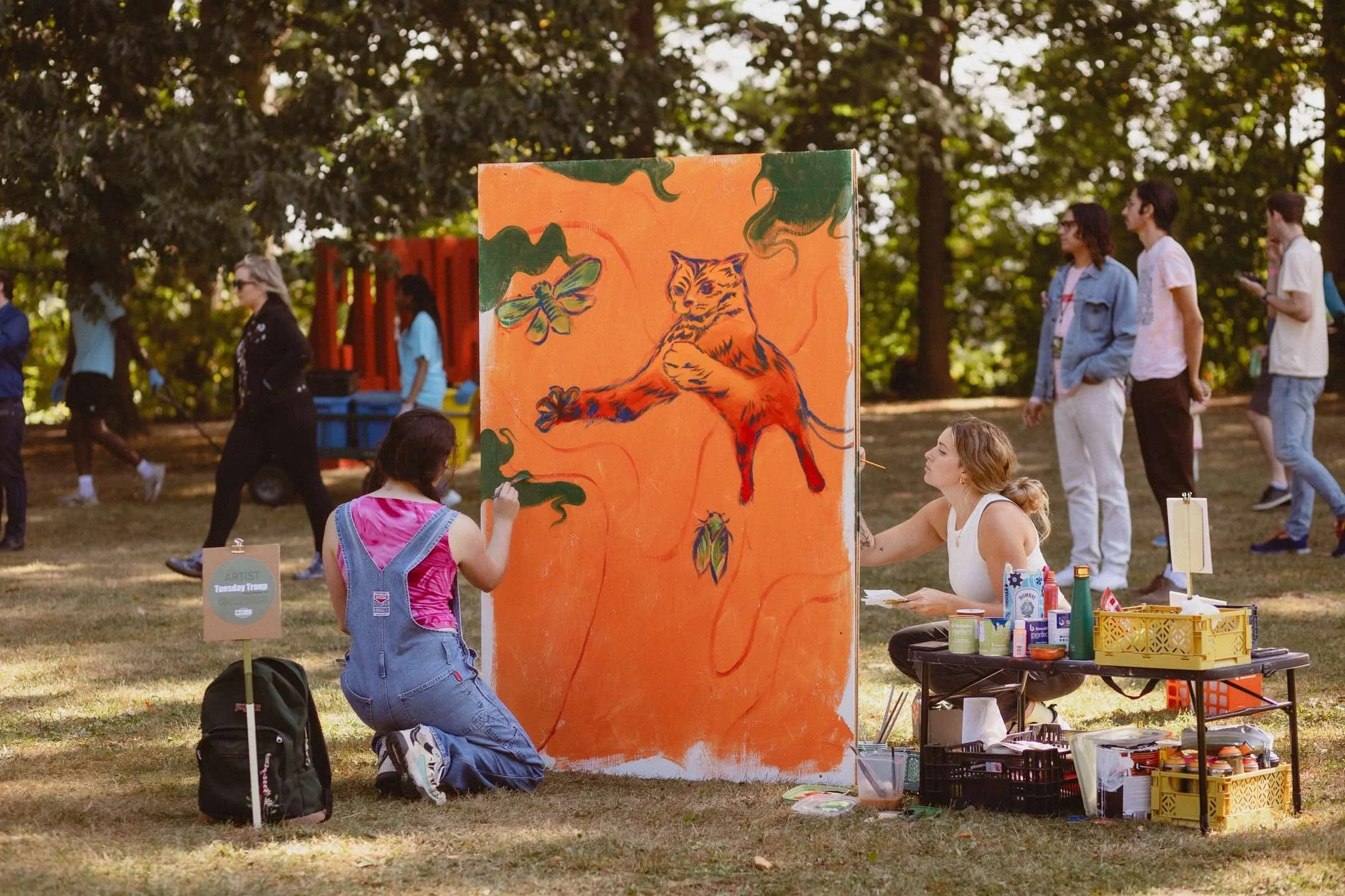 Two artists painting a colorful mural with a tiger on an orange background in a park setting, surrounded by trees and people in the background.