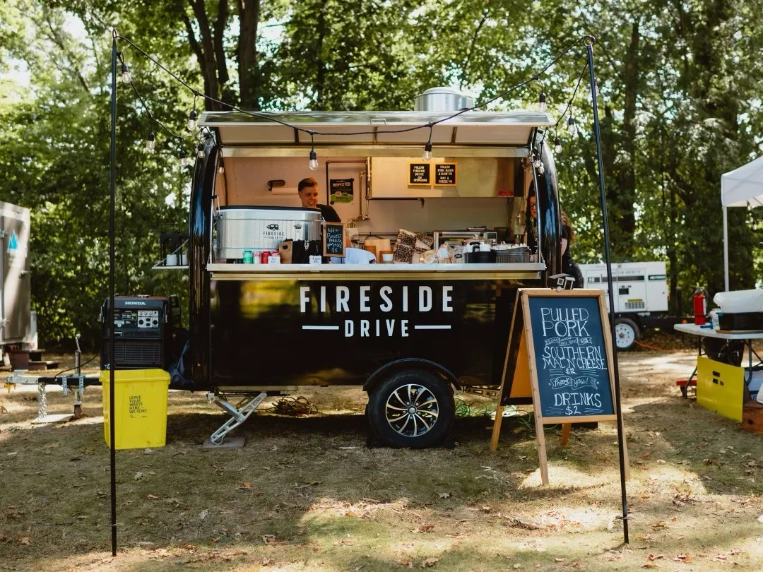 Food truck with "Fireside Drive" branding in a park setting, serving pulled pork and mac 'n' cheese from a chalkboard menu. Outdoor event with trees in the background.