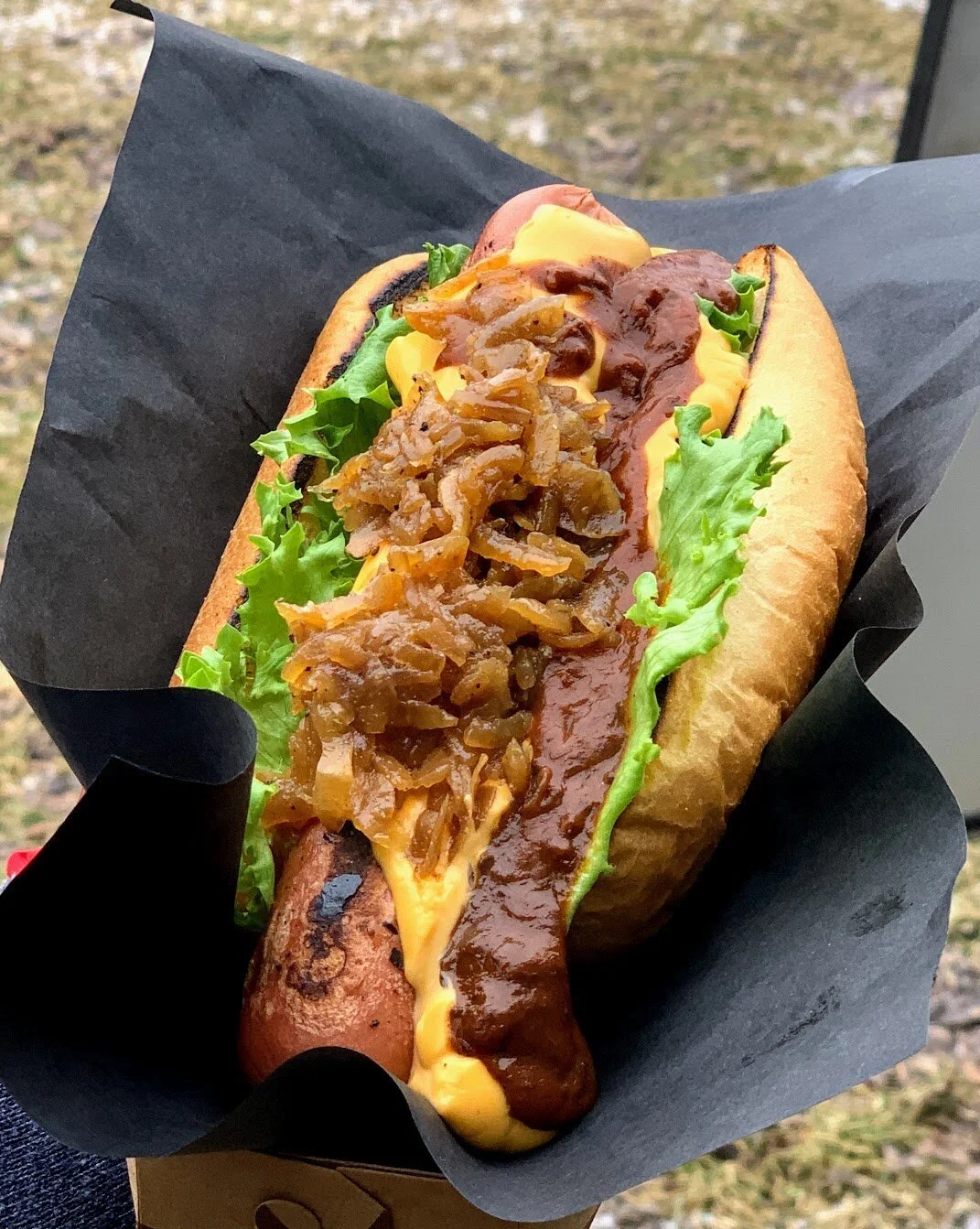 Close-up of a hot dog topped with melted cheese, chili, caramelized onions, and lettuce, wrapped in black paper.