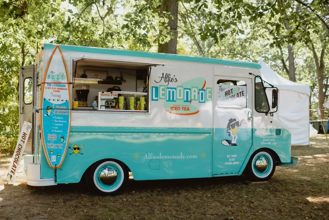 A vintage-style lemonade truck parked outdoors under trees, featuring colorful signage and a menu with various lemonade and iced tea options. The truck has a surfboard displaying the menu and serves items like hot chocolate. The brand name 'Alfie's Lemonade' is prominently displayed.