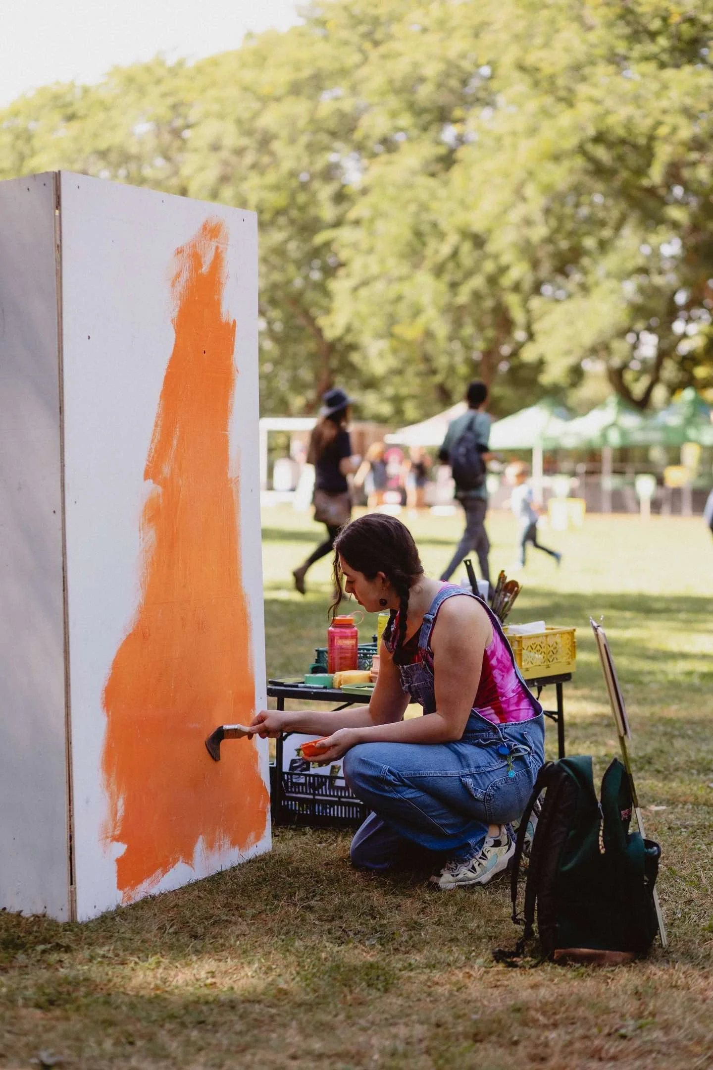 Artist painting an orange mural on a wooden panel outdoors in a park setting.