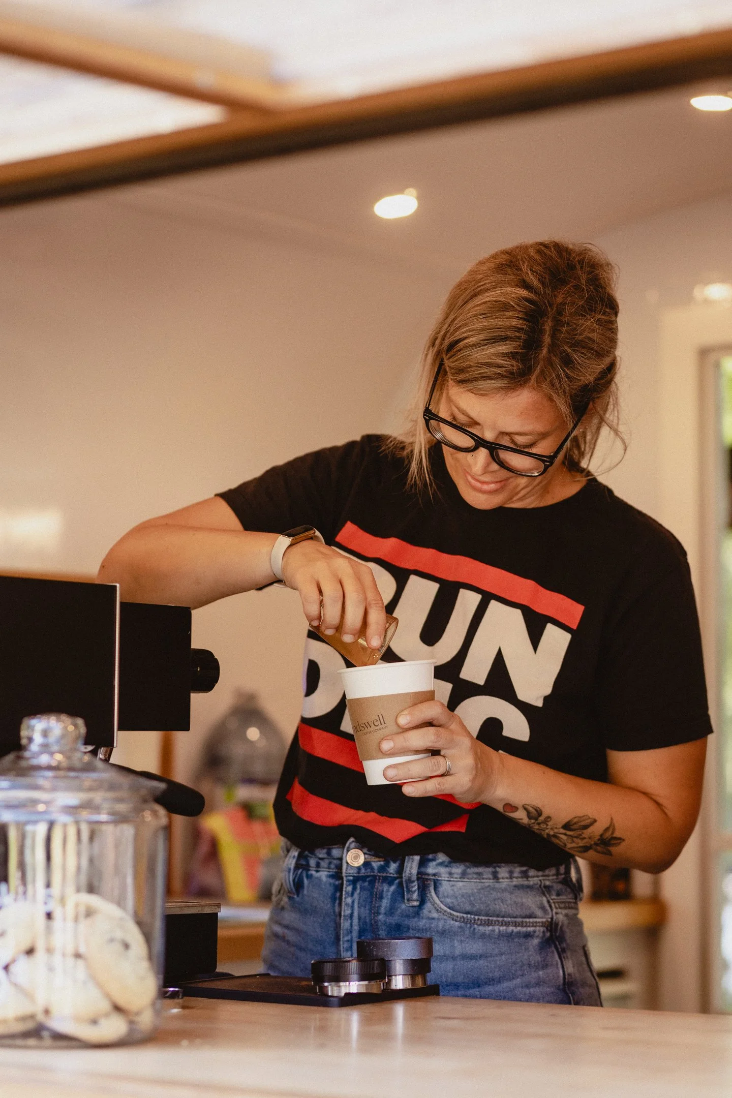Person wearing glasses and a "RUN DMC" shirt preparing a coffee drink in a cafe setting.
