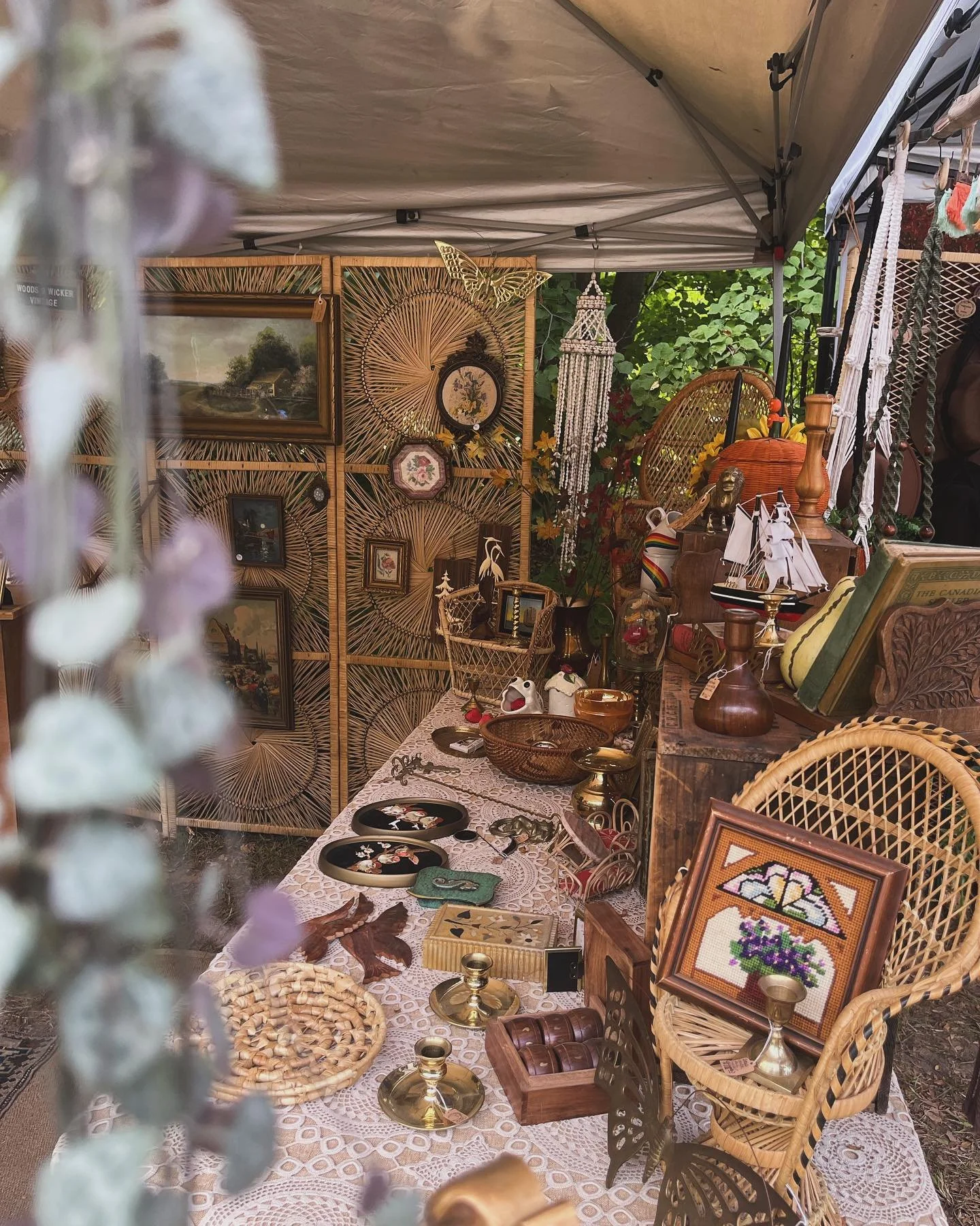 Outdoor vintage market stall with decorative items, wicker furniture, and framed pictures. Displayed on a lace-covered table are various trinkets, a model ship, candlesticks, and woven baskets. A macramé hanging decorates the background.