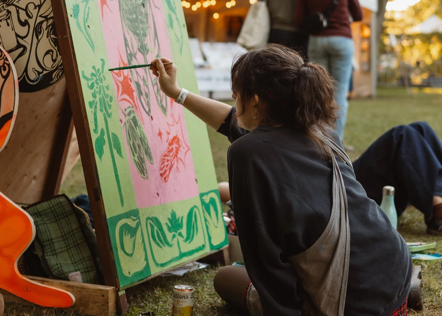 Person painting a colorful mural on a wooden board outdoors, with a beverage can and people in the background.