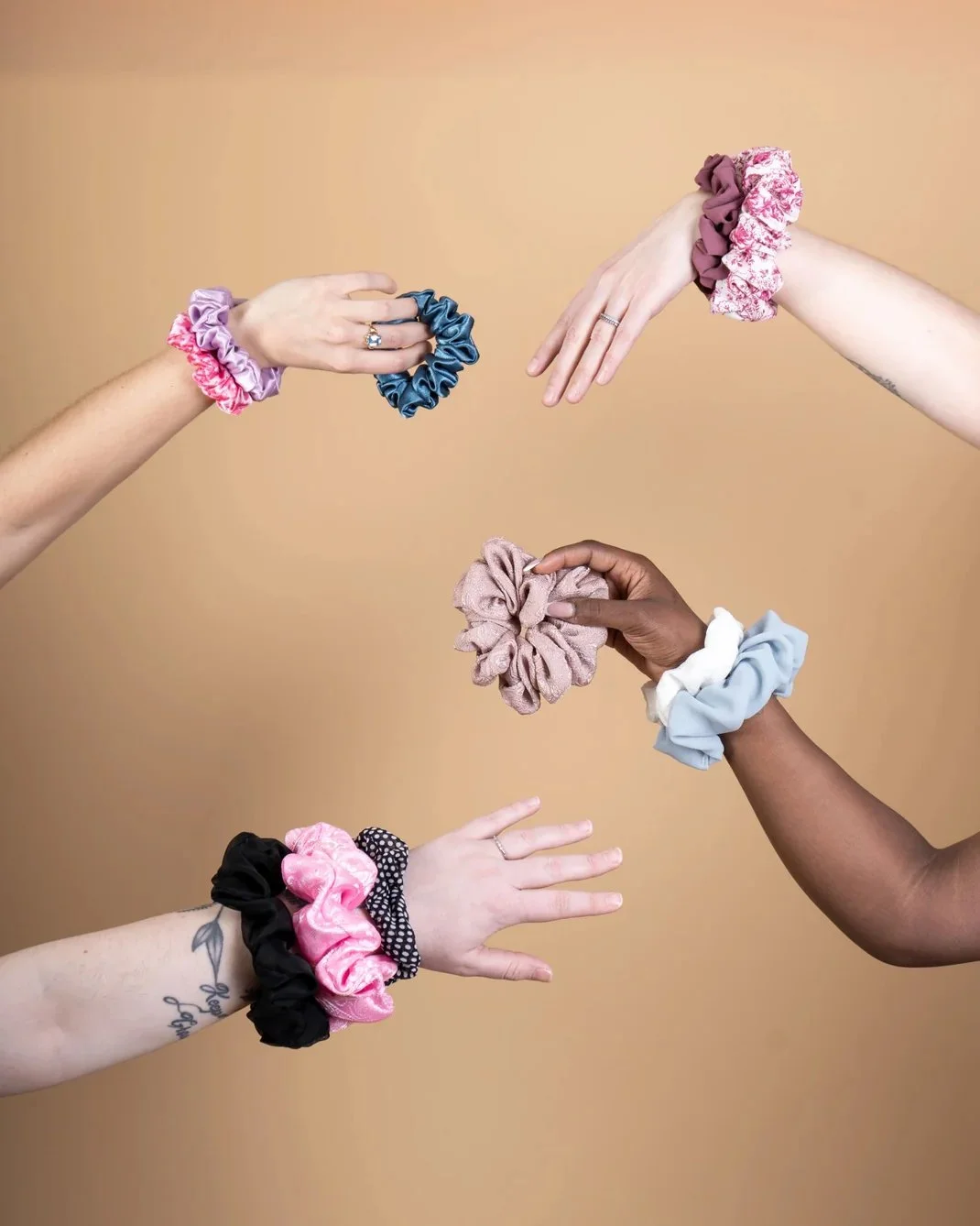 Various hands holding colorful scrunchies against a beige background.