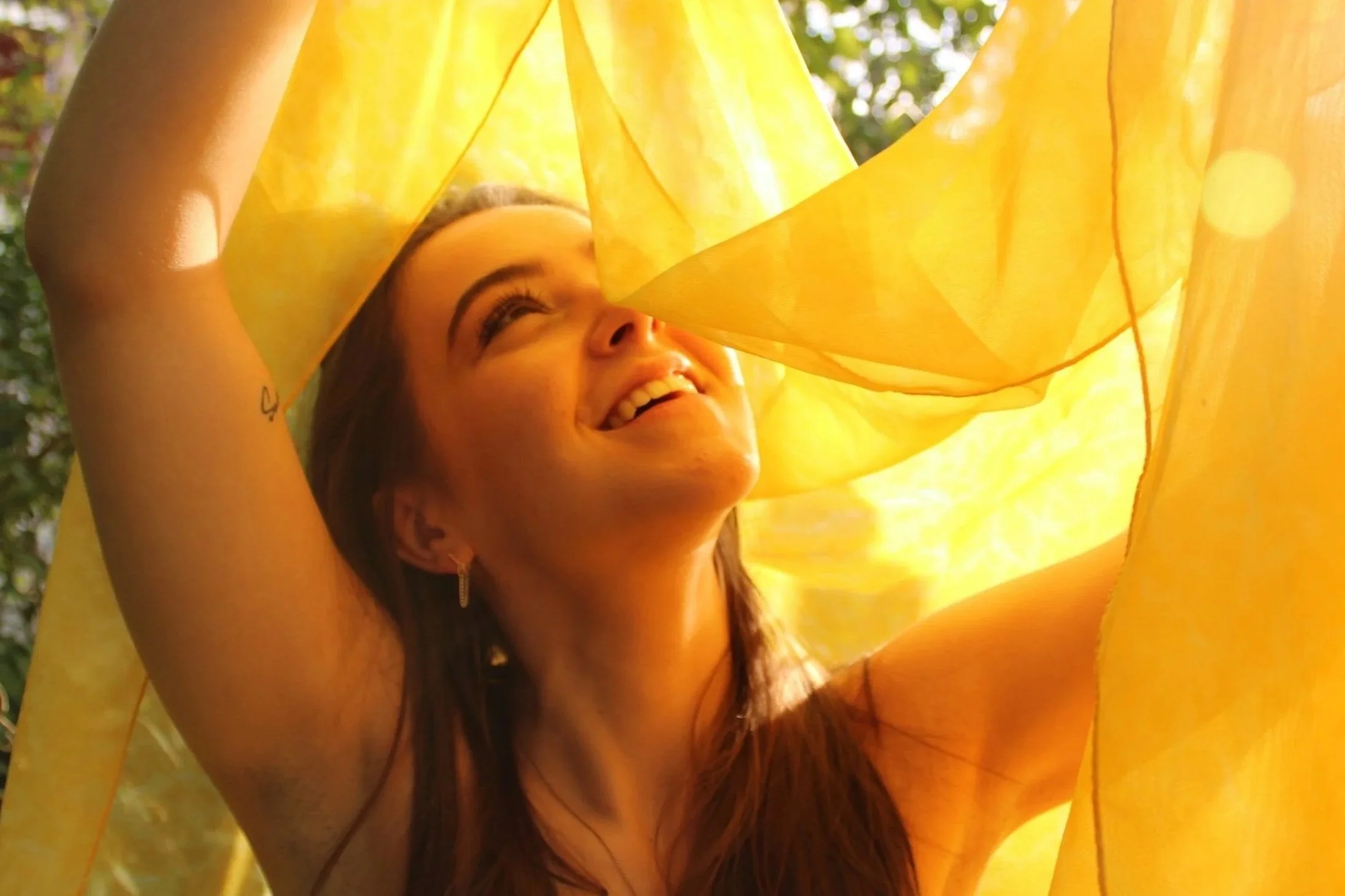 A young woman smiling and looking up, holding a yellow fabric or curtain above her head, with sunlight creating a warm, glowing effect.
