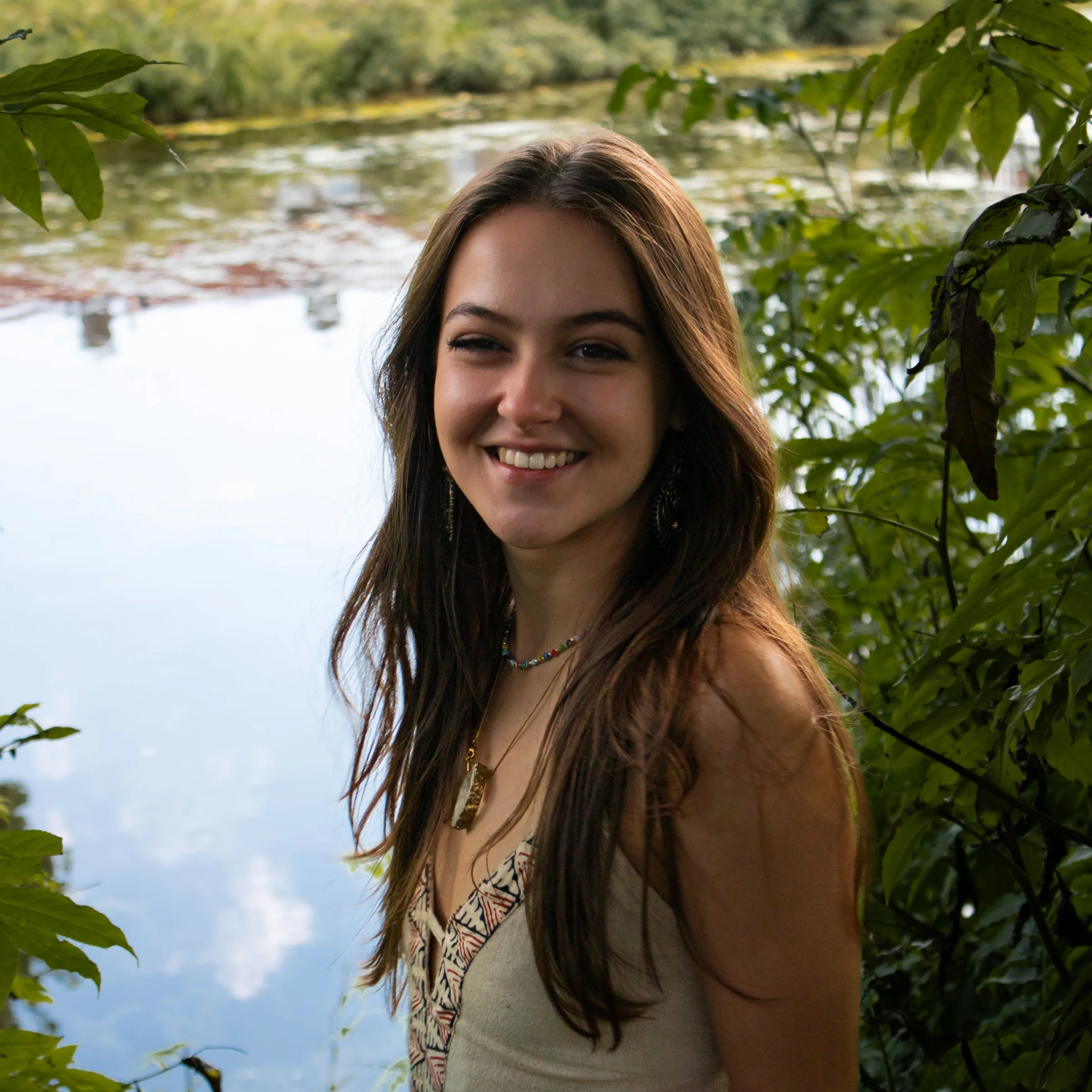 A young woman with long brown hair smiling outdoors near a body of water, surrounded by green foliage.