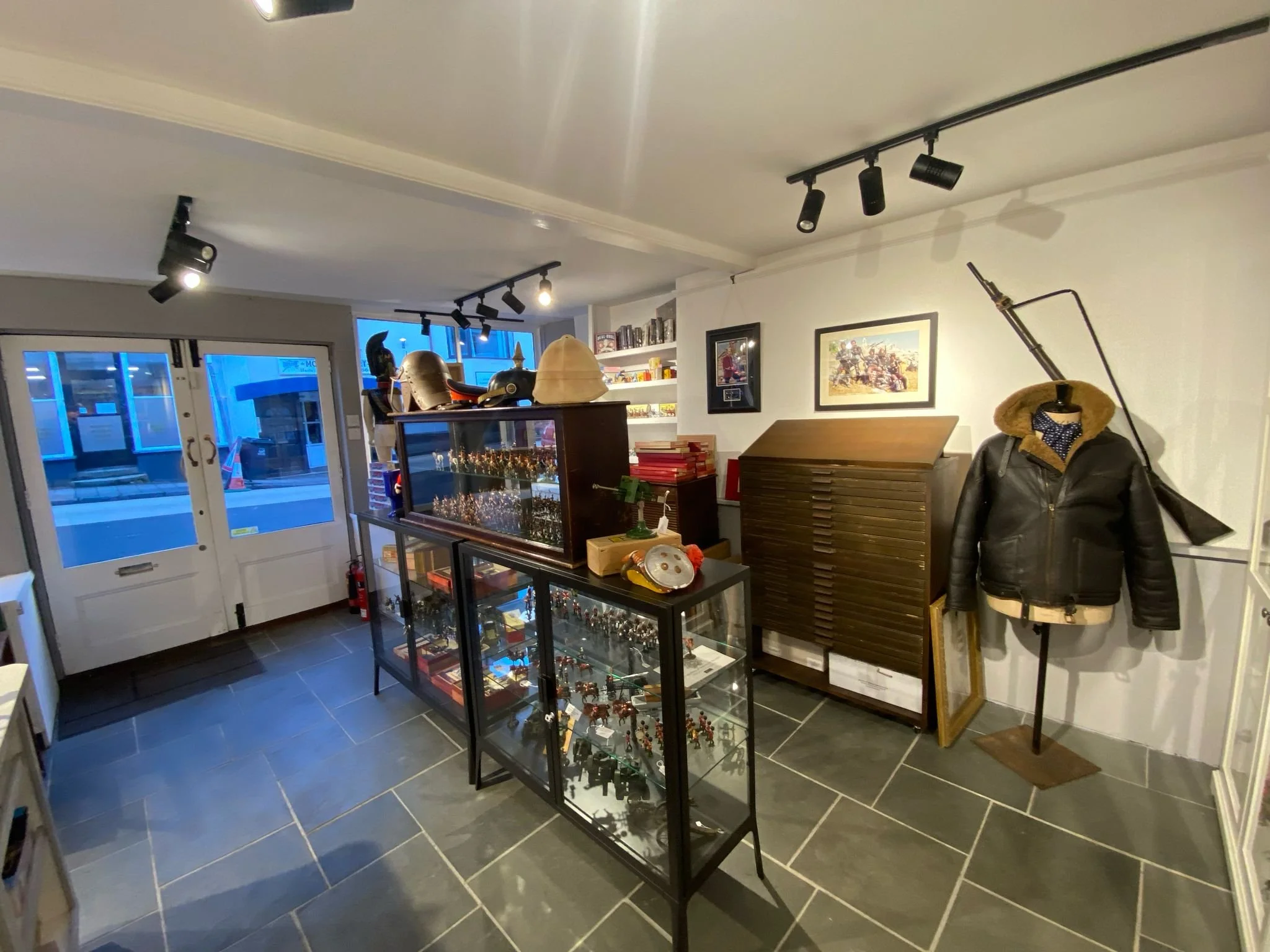 Interior of a store with glass display cases filled with collectibles, a wall-mounted mannequin dressed in a leather jacket with a helmet, and a coat rack with helmets and a large gun prop mounted on the wall, illuminated by ceiling spotlights.