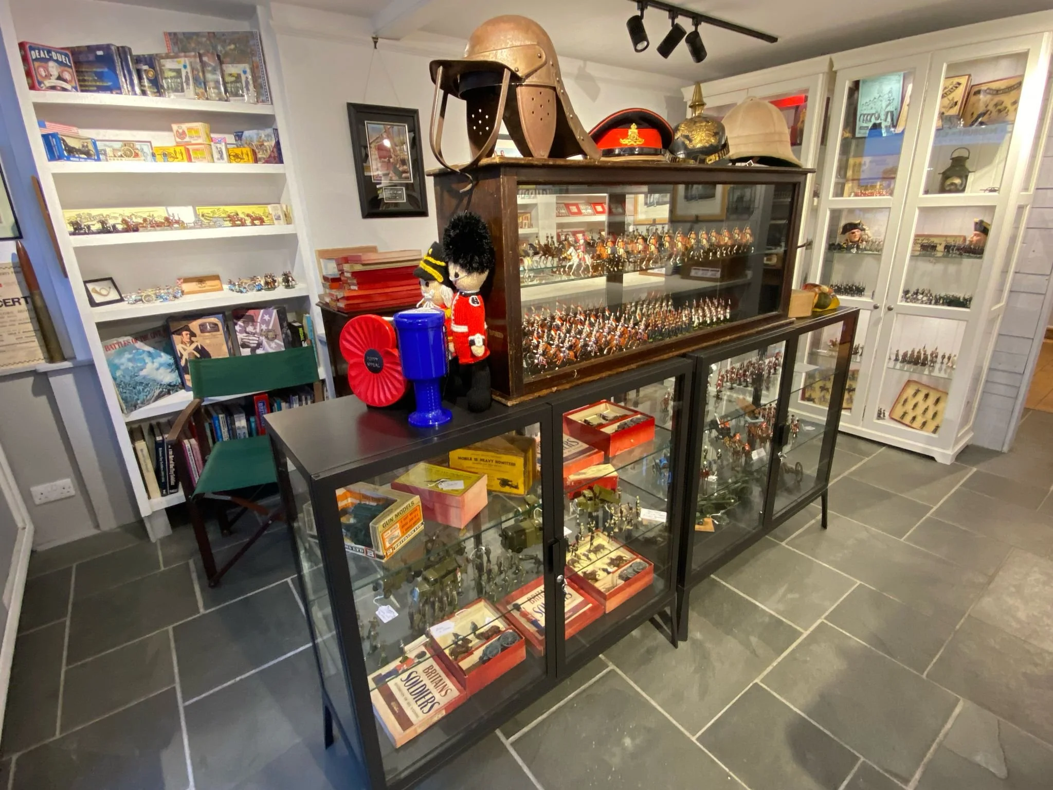 Room with display cabinets filled with miniature soldiers, military helmets, and collectibles. Shelves on the left hold board games and books. A small green chair and various military memorabilia are present.