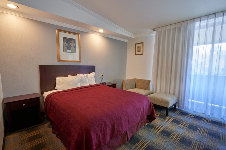 Hotel room with a double bed featuring a red bedspread, two wooden nightstands, abstract artwork on the walls, and a lounge chair by sheer curtains covering a window.