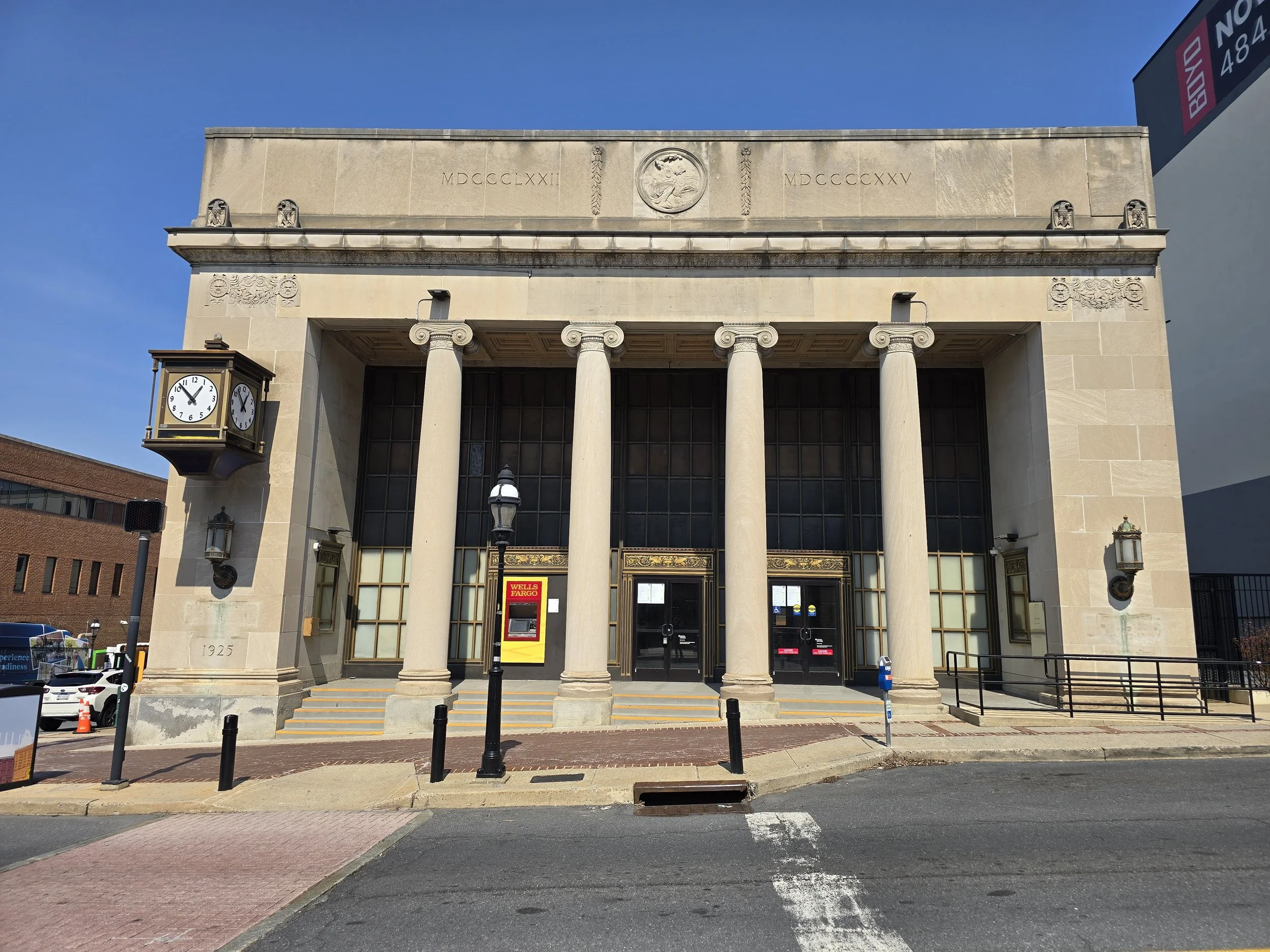 A historic-style building with four large columns in front, an ornate clock on the left side, and a bank ATM in the center. The building has Roman numerals and a round emblem on the upper facade, with a clear blue sky overhead.
