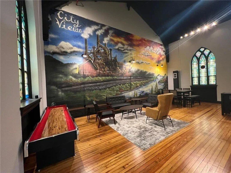 A cozy lounge with wooden floors, a colorful mural of a city and train tracks on the wall, vintage furniture including a yellow armchair and black sofas, a shuffleboard table, and arched stained glass windows.