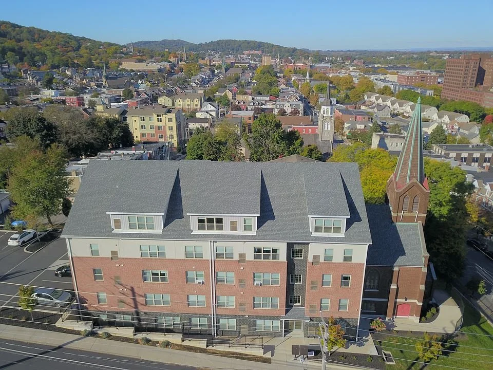 A large multi-story brick building with a steep gray roof and dormer windows, adjacent to a church with a tall steeple, located in an urban area with residential buildings and trees, under a clear blue sky.