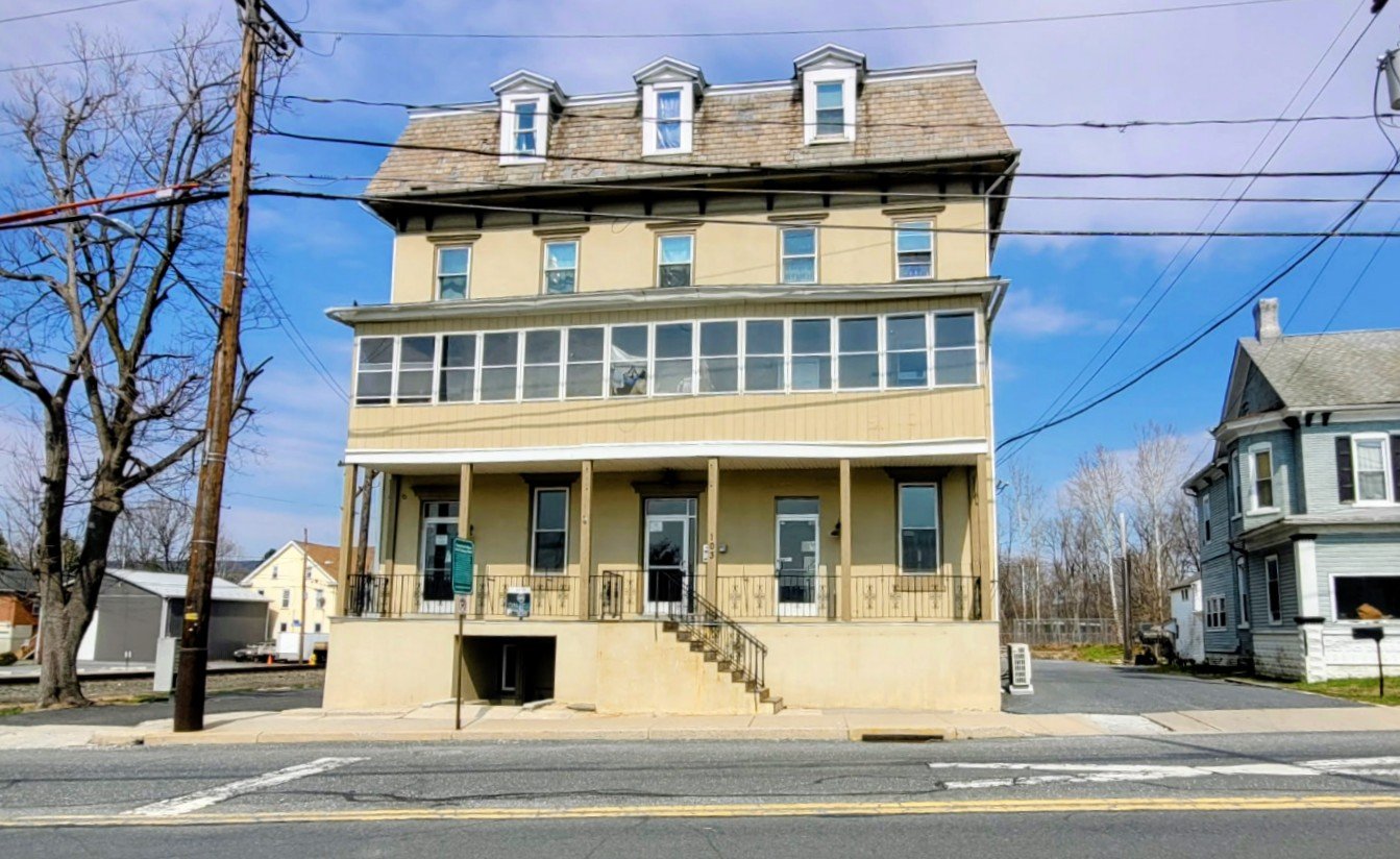 Four-story beige house with a steep roof and multiple dormer windows, situated on a street, with visible power lines and a blue sky.