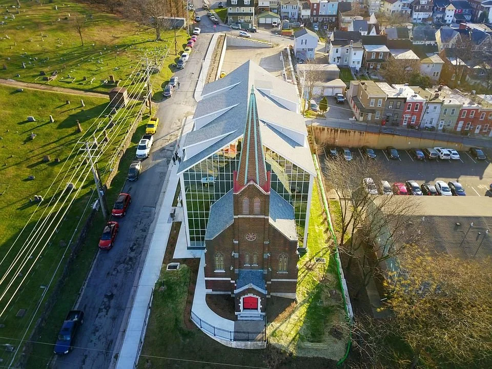 An aerial view of a church with a brick facade, tall steeple, and large glass windows, situated between a parking lot and a residential neighborhood in an urban area.