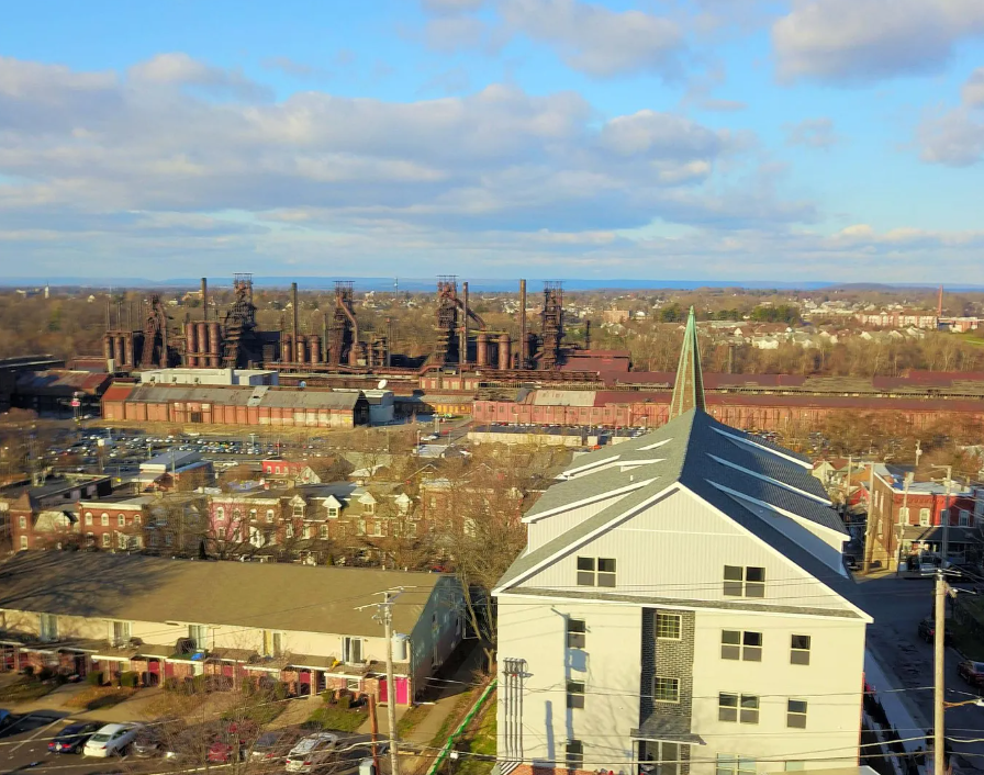 A cityscape with a white church in the foreground, a large industrial building with smokestacks in the middle, and a horizon of buildings and trees under a partly cloudy sky.