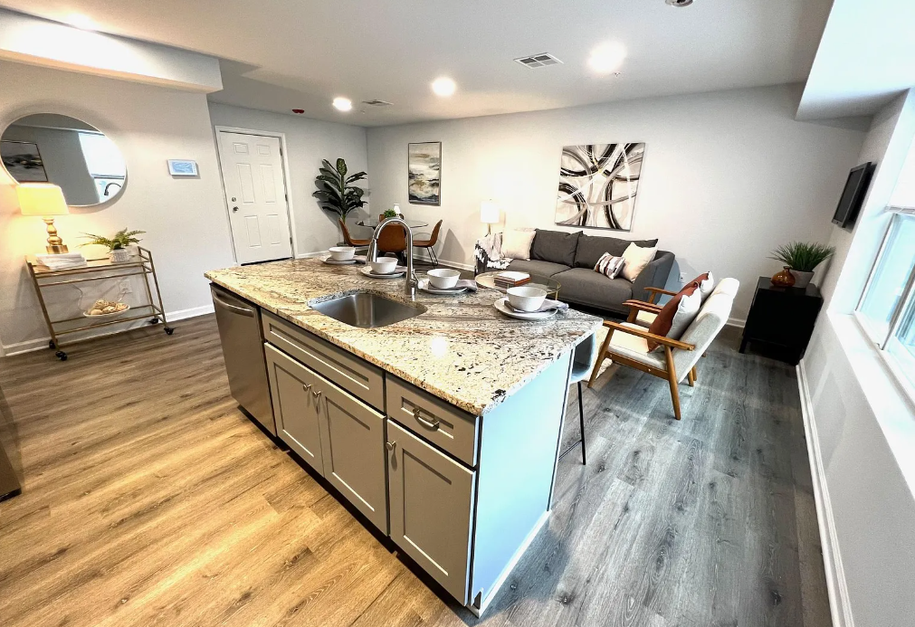 Open-concept living room and kitchen with granite island, gray sofa, and wooden chairs, decorated in neutral tones.
