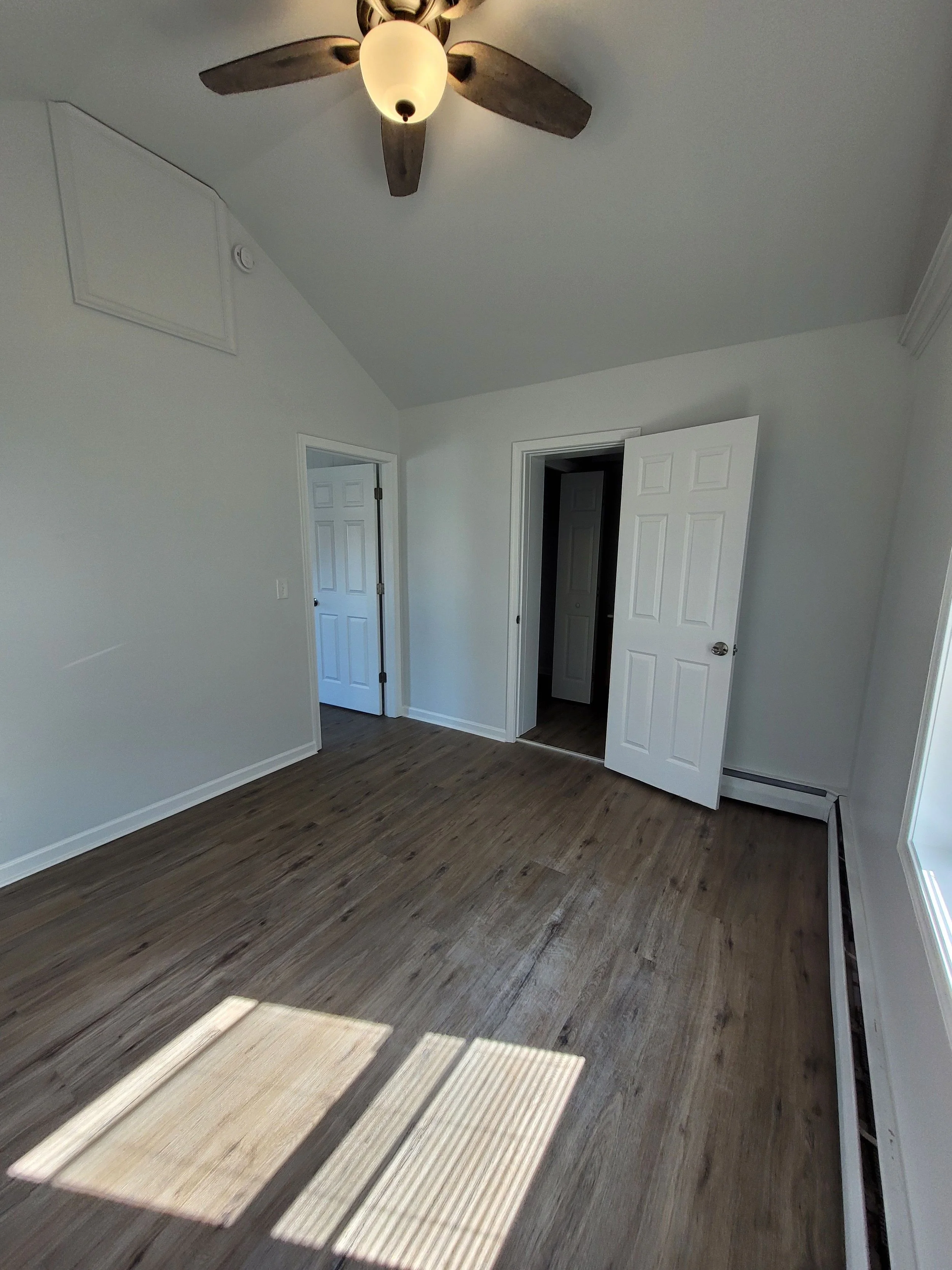 Empty room with wooden floors, white walls, an open door leading to another room, a closed door, a ceiling fan with light, and a large window letting in natural light.