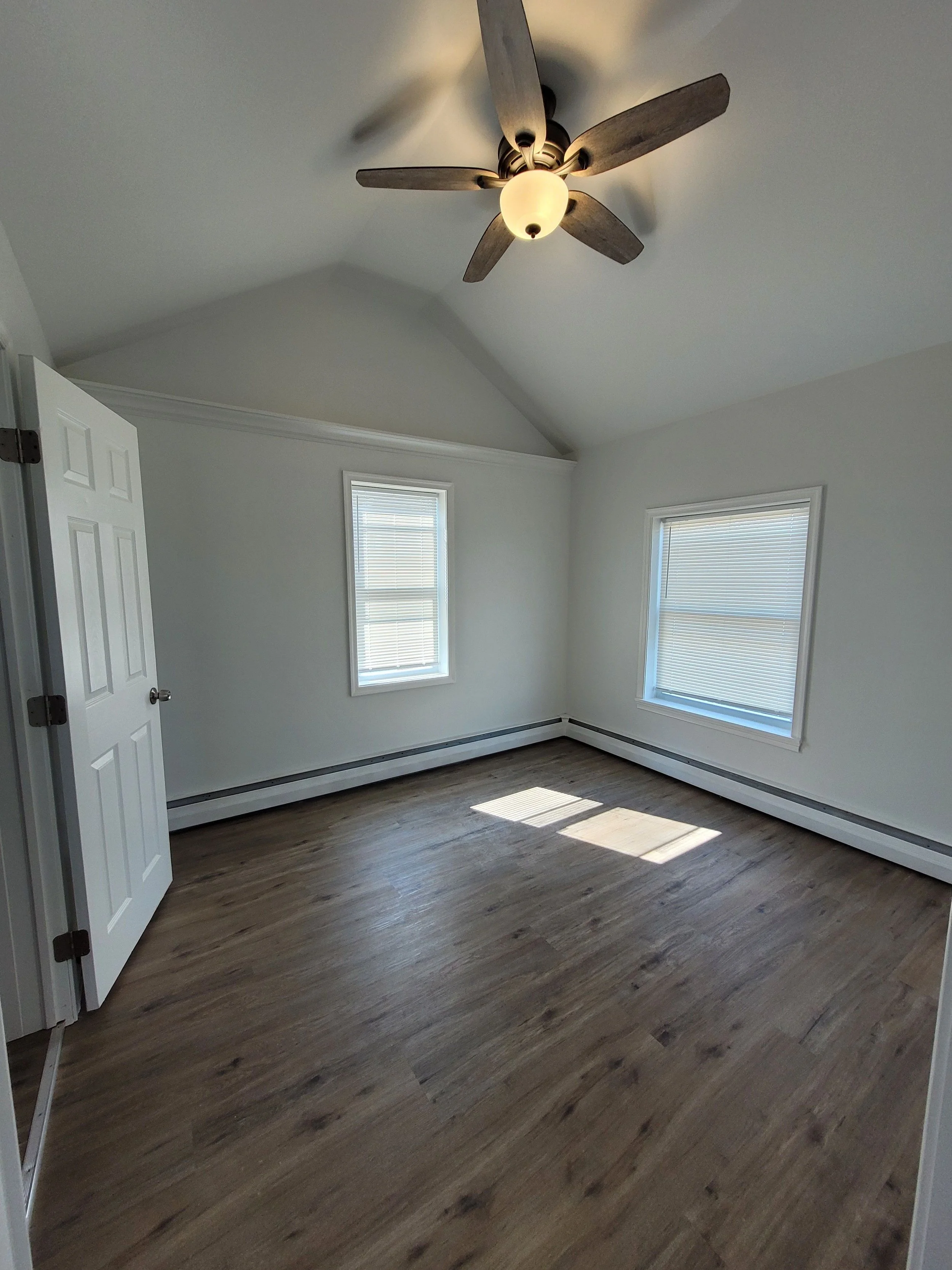 Empty room with white walls, two windows with blinds, a ceiling fan with lights, and wood flooring.