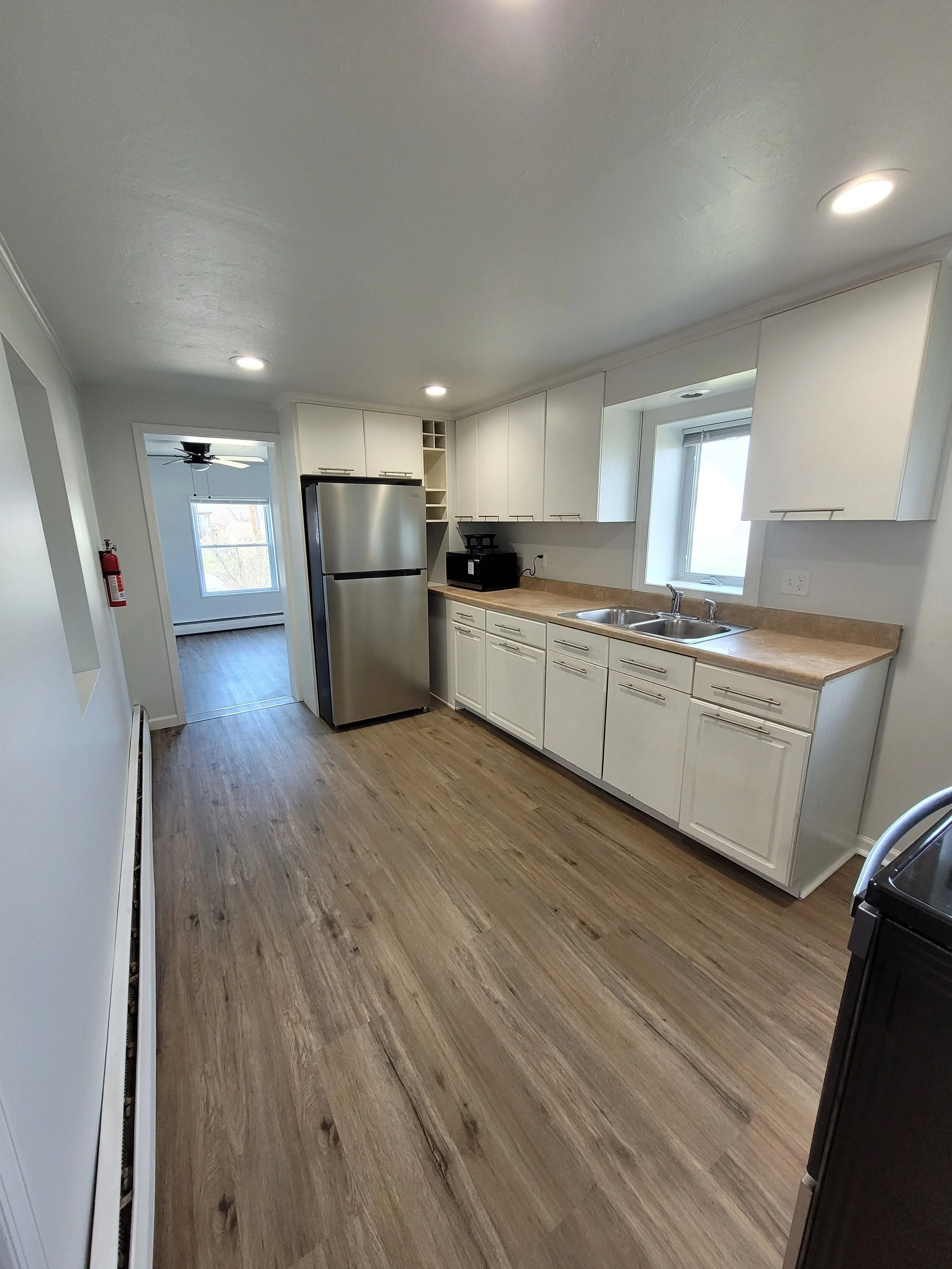 Clean, modern kitchen with white cabinets, beige countertop, stainless steel refrigerator, double sink, and wooden flooring. There are recessed ceiling lights and a view into an adjacent room with a ceiling fan and window.