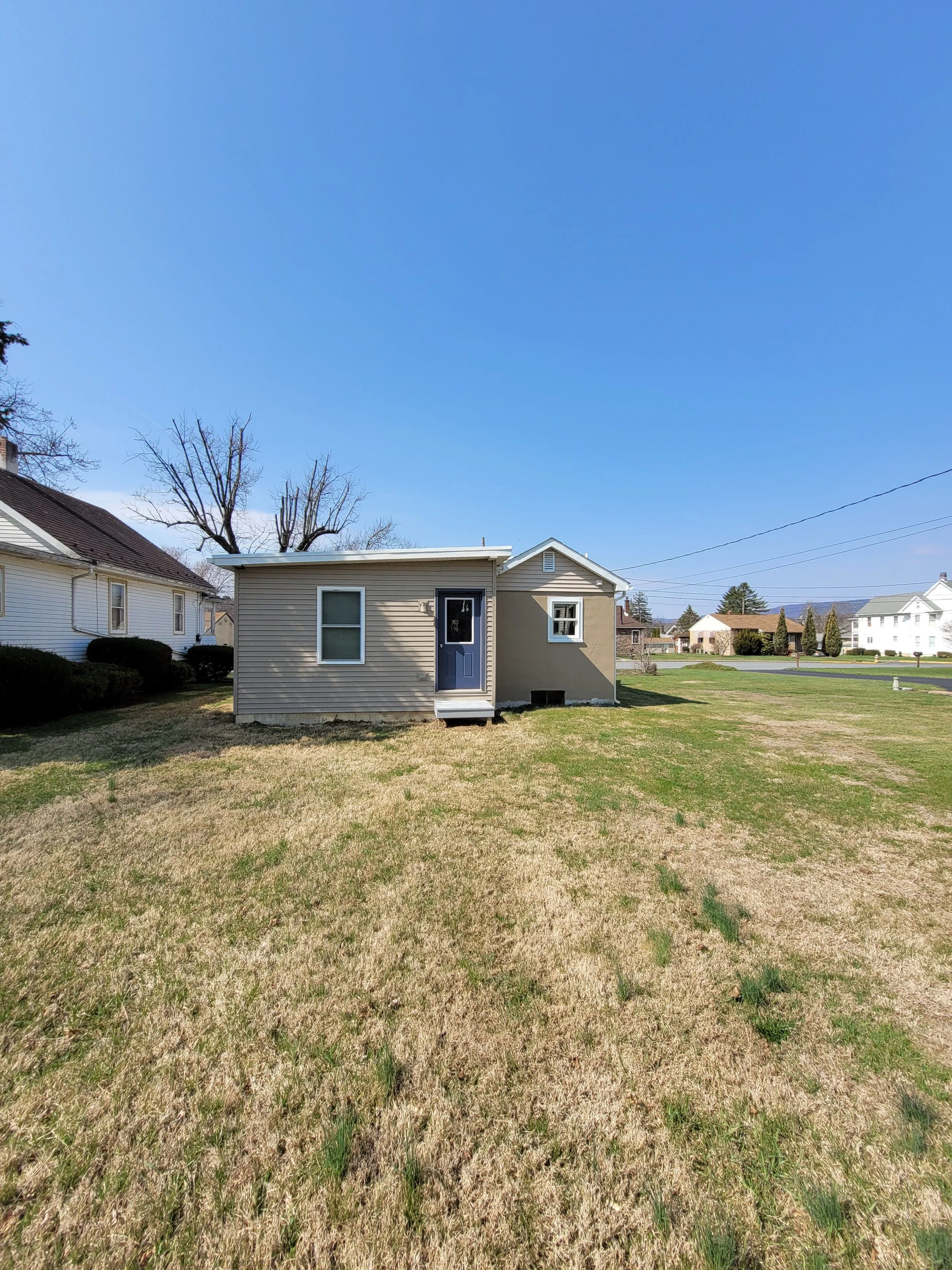 Small beige house with a blue door, small front steps, on a grassy lawn under a clear blue sky, neighboring houses in the background.