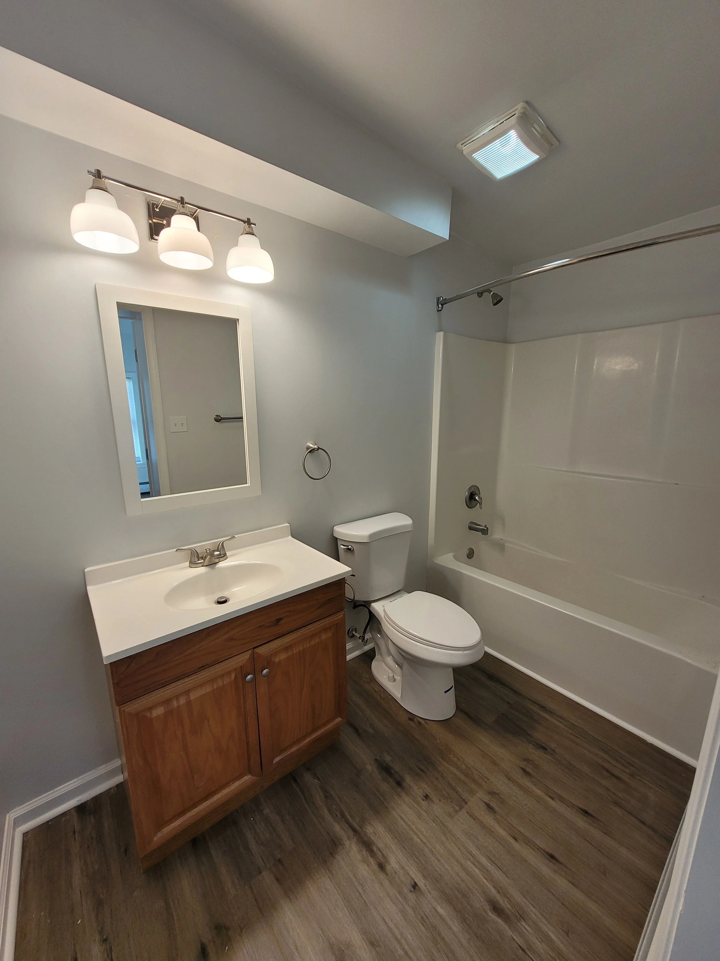 A bathroom with a mirror, vanity with a sink, toilet, bathtub, and wood flooring.