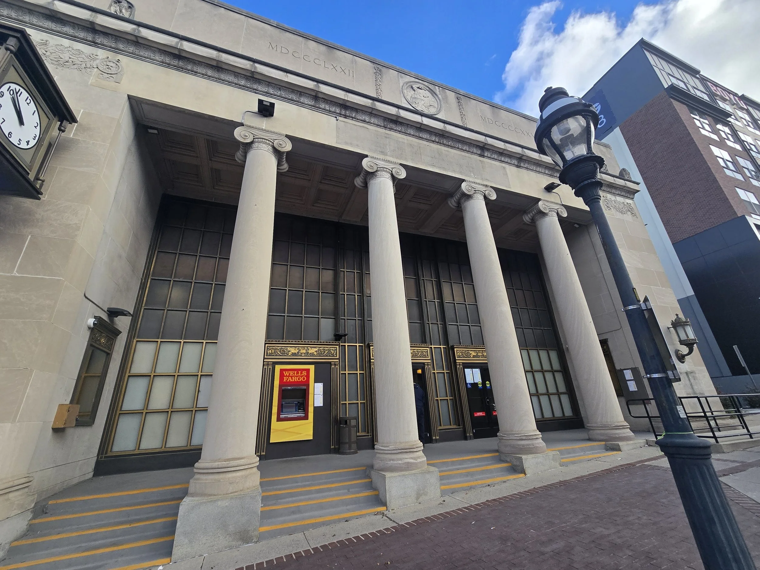 Exterior of a classical-style building with large columns, intricate carvings, and a clock on the wall. A Wells Fargo bank ATM is visible near the entrance. A street lamp leans towards the building, and the sky is partly cloudy.